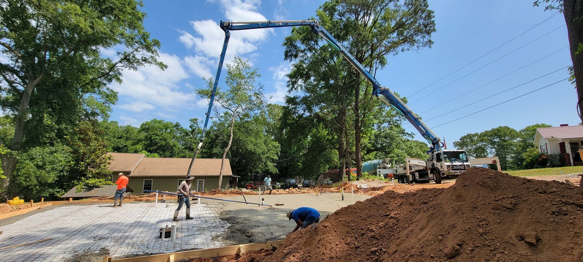 Construction site with workers pouring concrete beside a dirt mound and trees under a blue sky