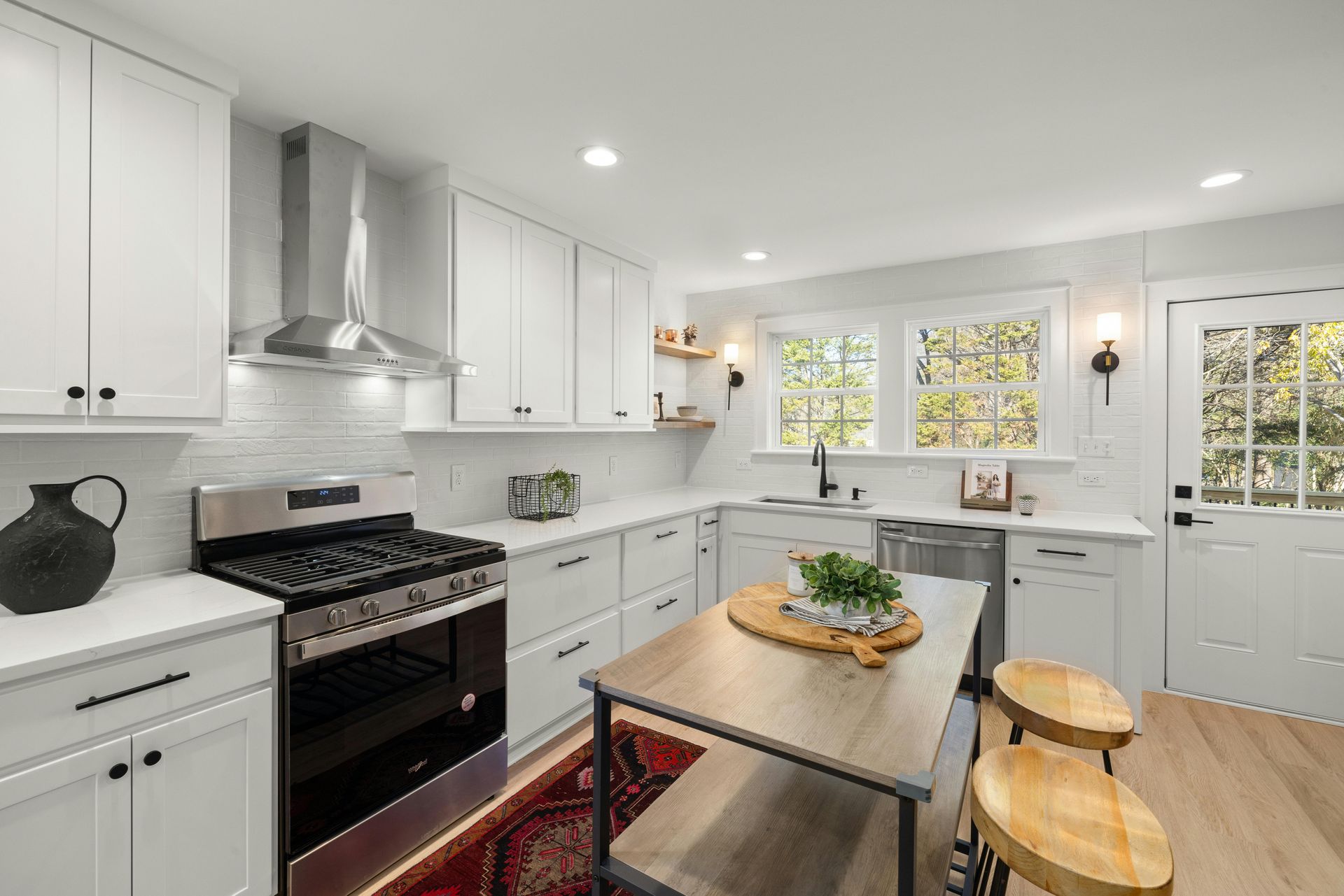 Bright white kitchen with black stove, island table, and stools, lit by windows and recessed ceiling lights