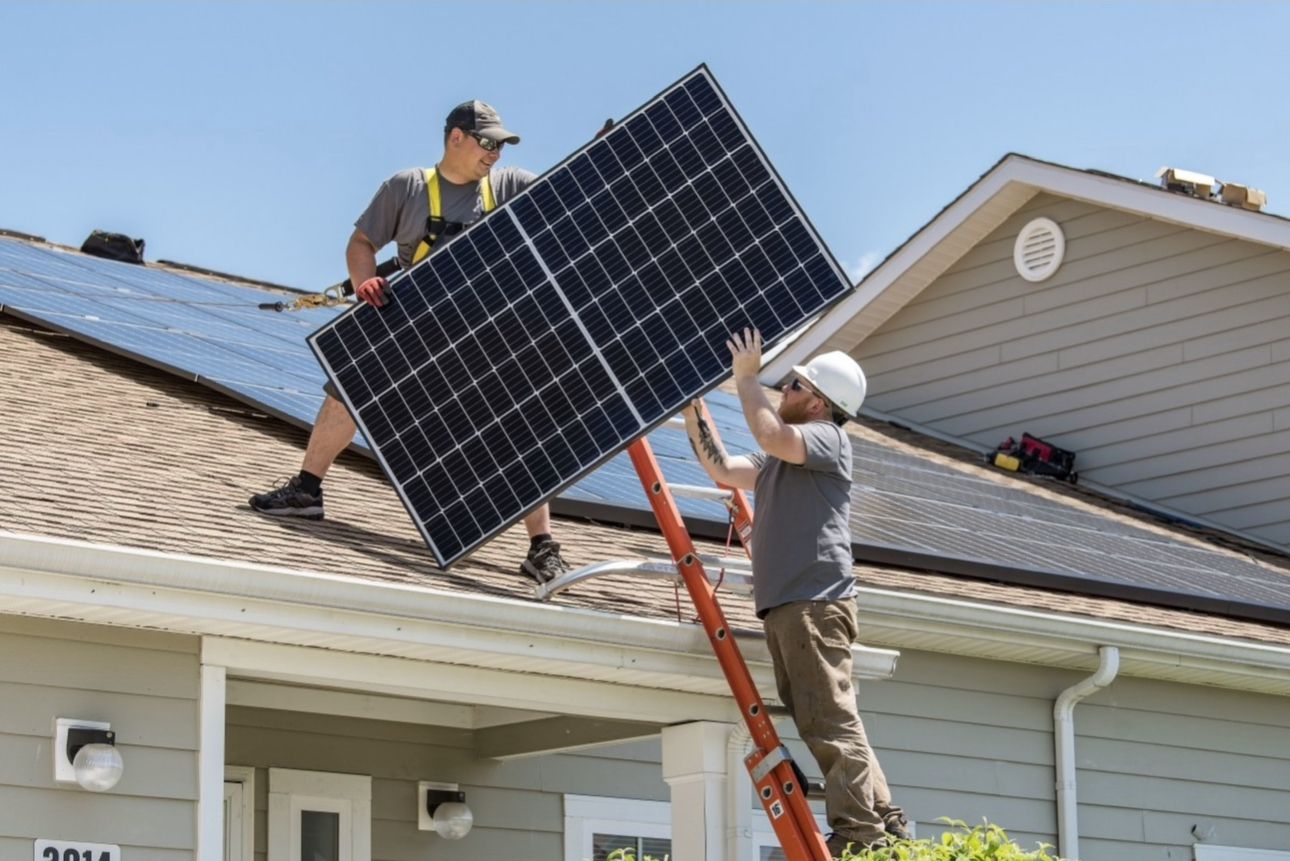 Two workers install a solar panel on a rooftop with a ladder.