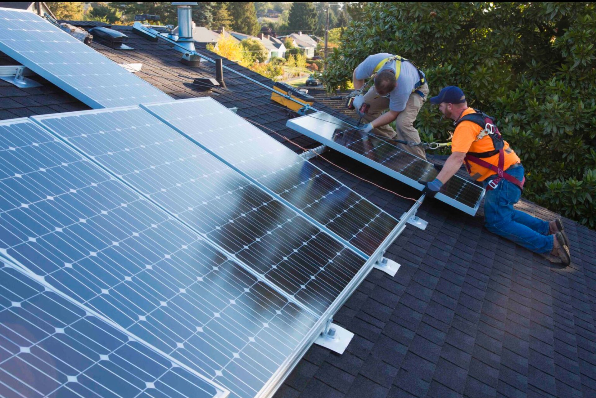 Workers installing solar panels on a shingled roof, one kneeling and one standing near the panels.