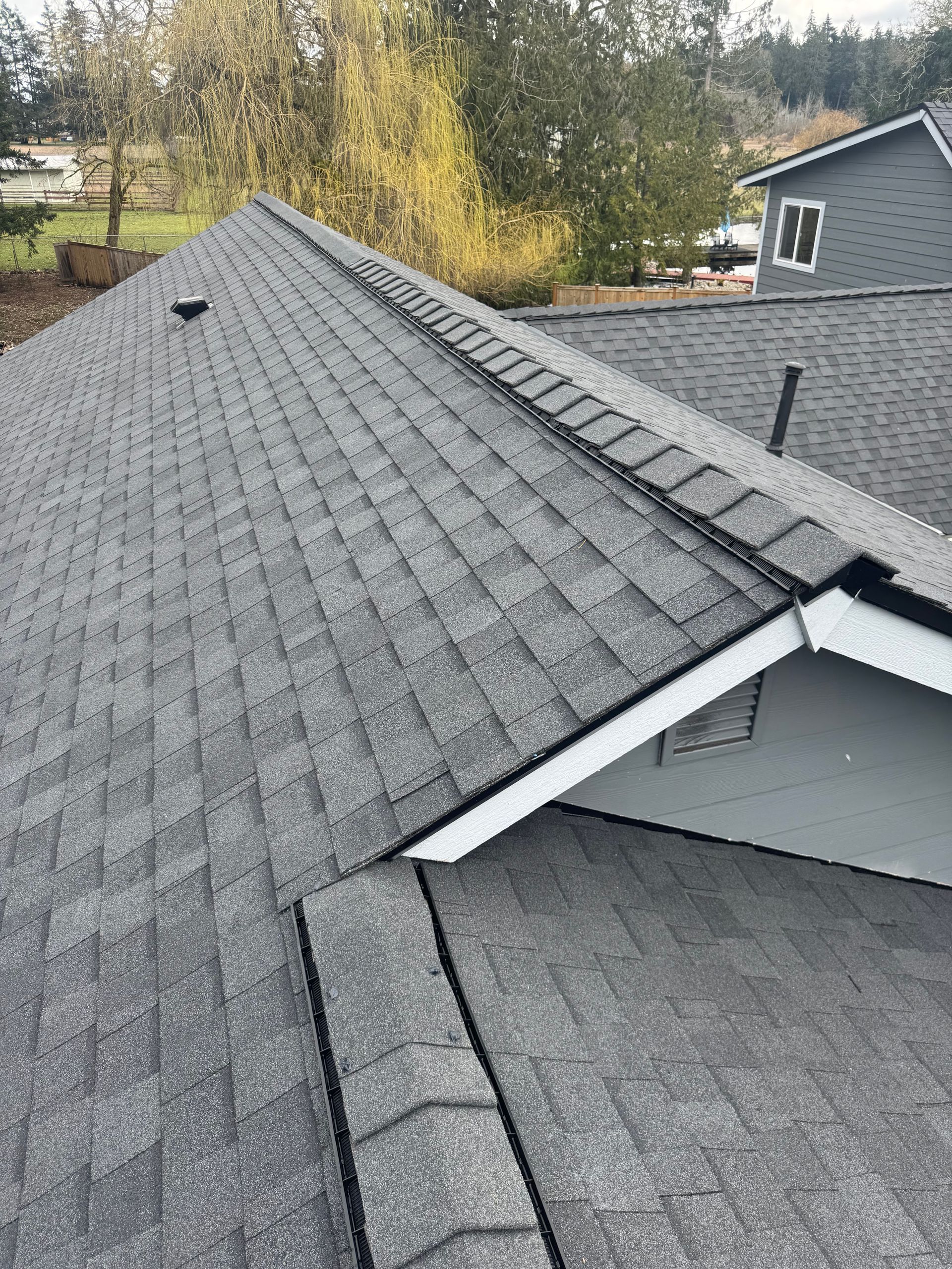 Gray shingled roof with a ridge vent and dark vent pipe, viewed from above near trees and a neighboring house
