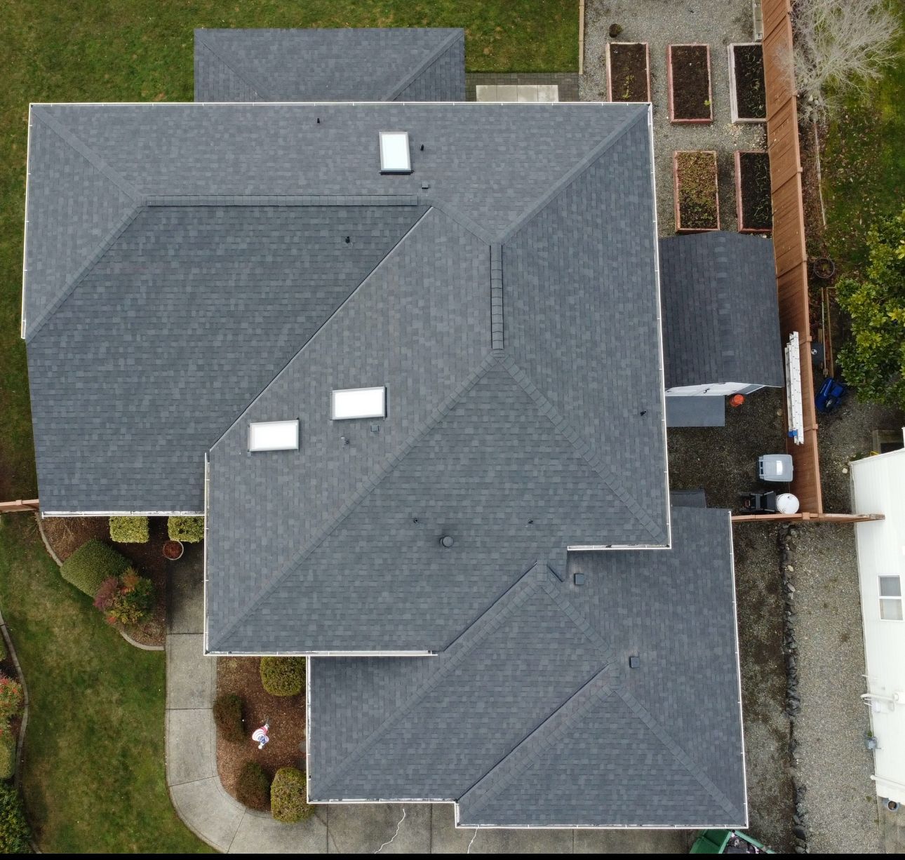 Aerial view of a gray-shingled house roof with skylights, surrounded by yard and neighboring buildings.
