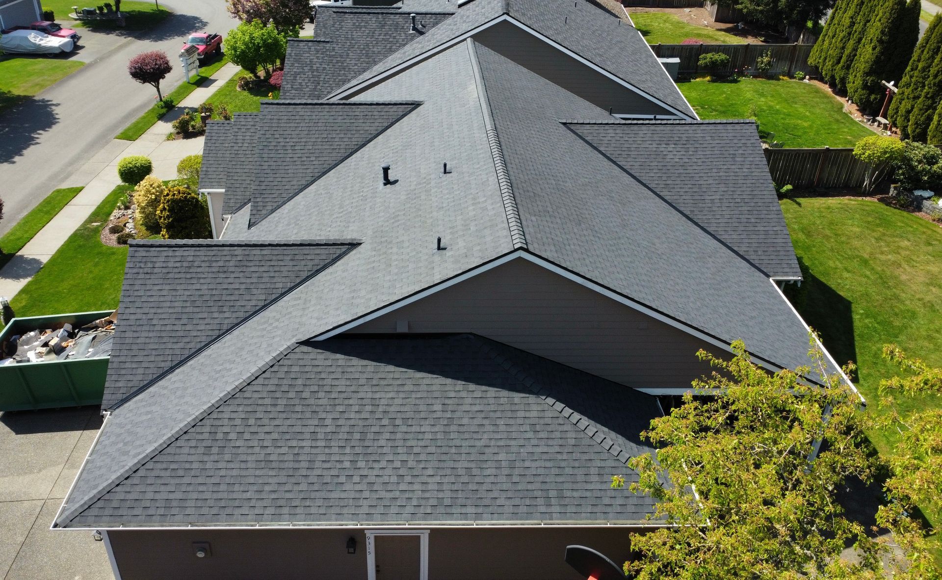 Aerial view of a gray shingled house roof in a suburban neighborhood with trees and parked cars nearby
