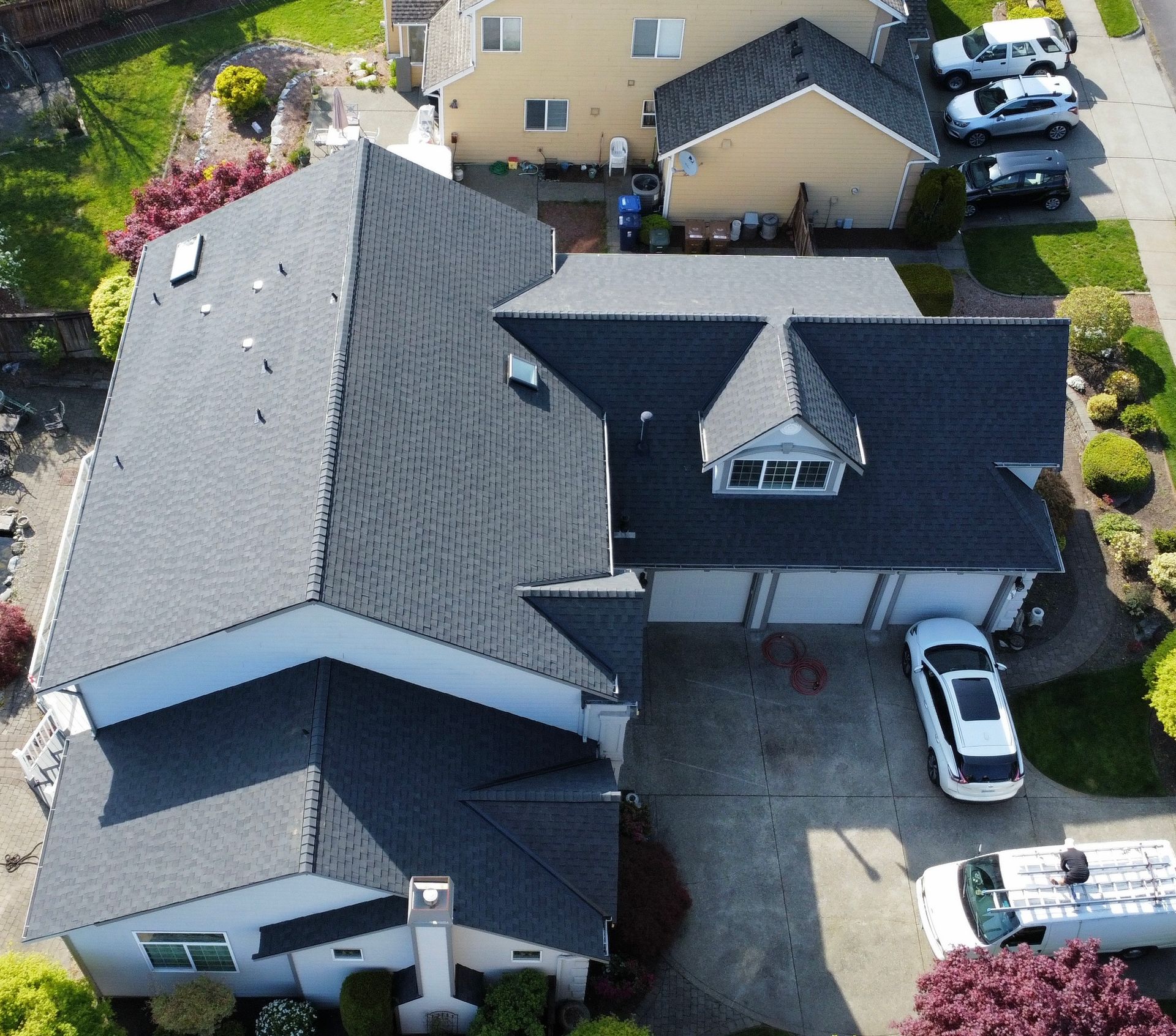 Aerial view of a suburban house with dark gray roofs, driveway, and parked cars.