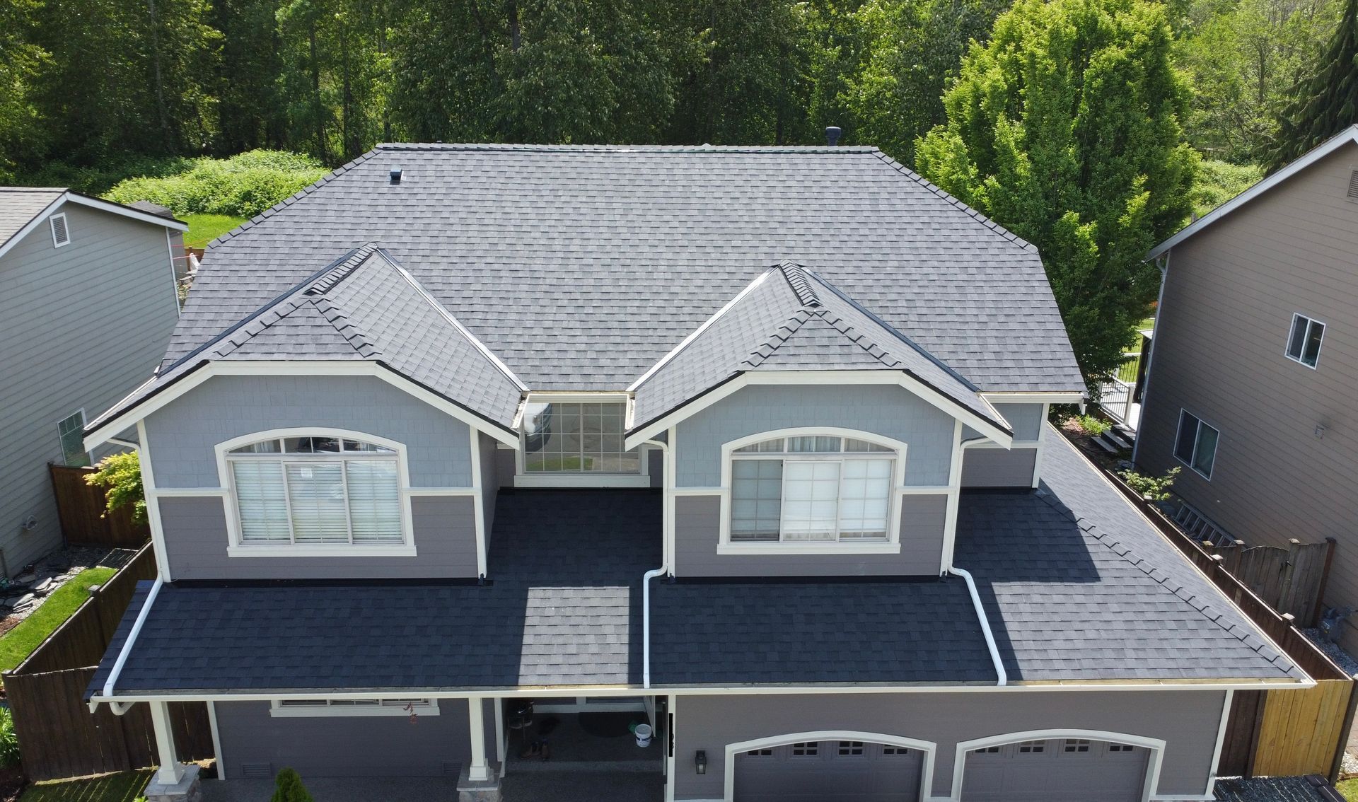 Aerial view of a gray-roofed suburban house with dormers and white trim, surrounded by trees.