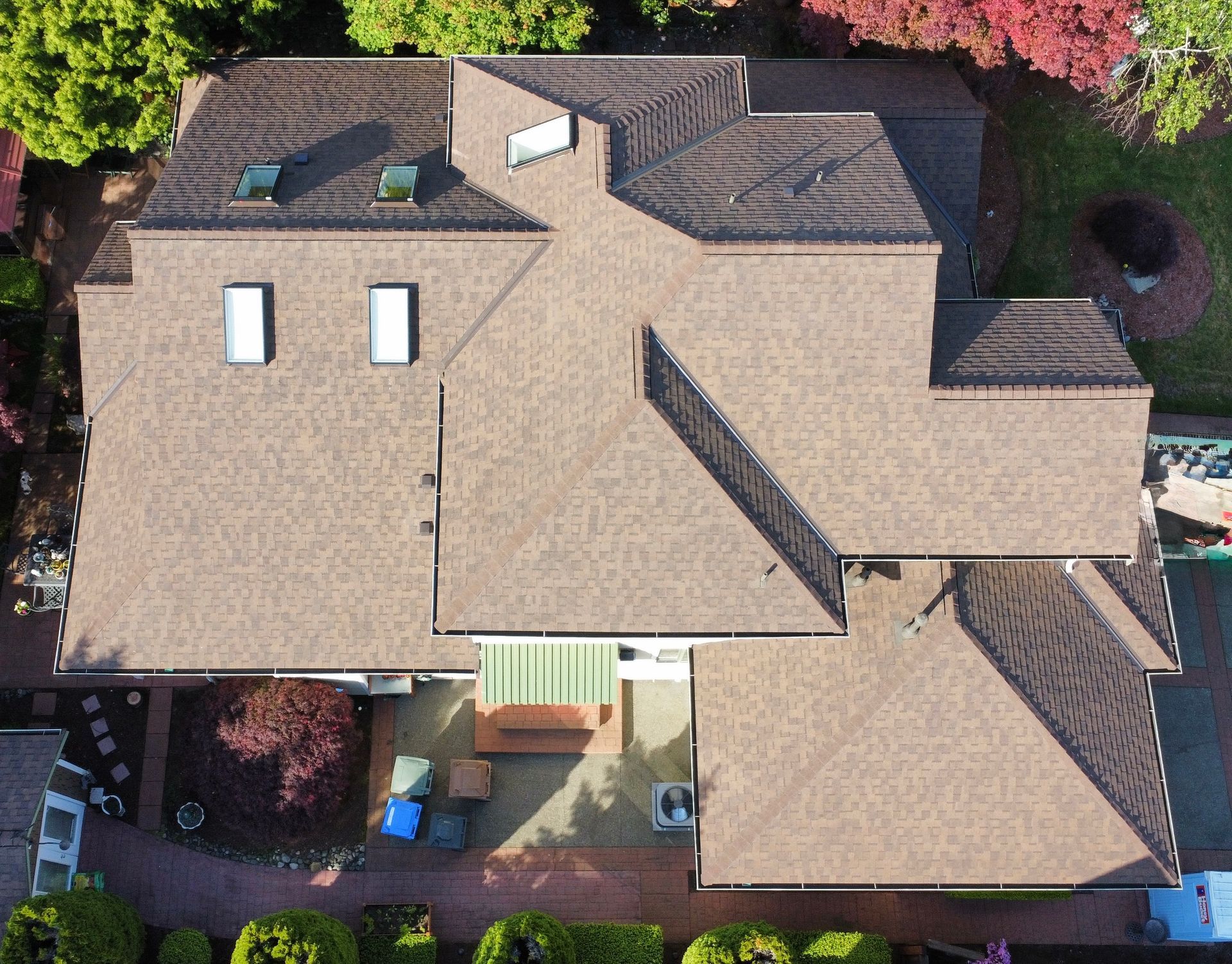 Aerial view of a tan-roofed house with skylights, surrounded by trees and a small backyard patio