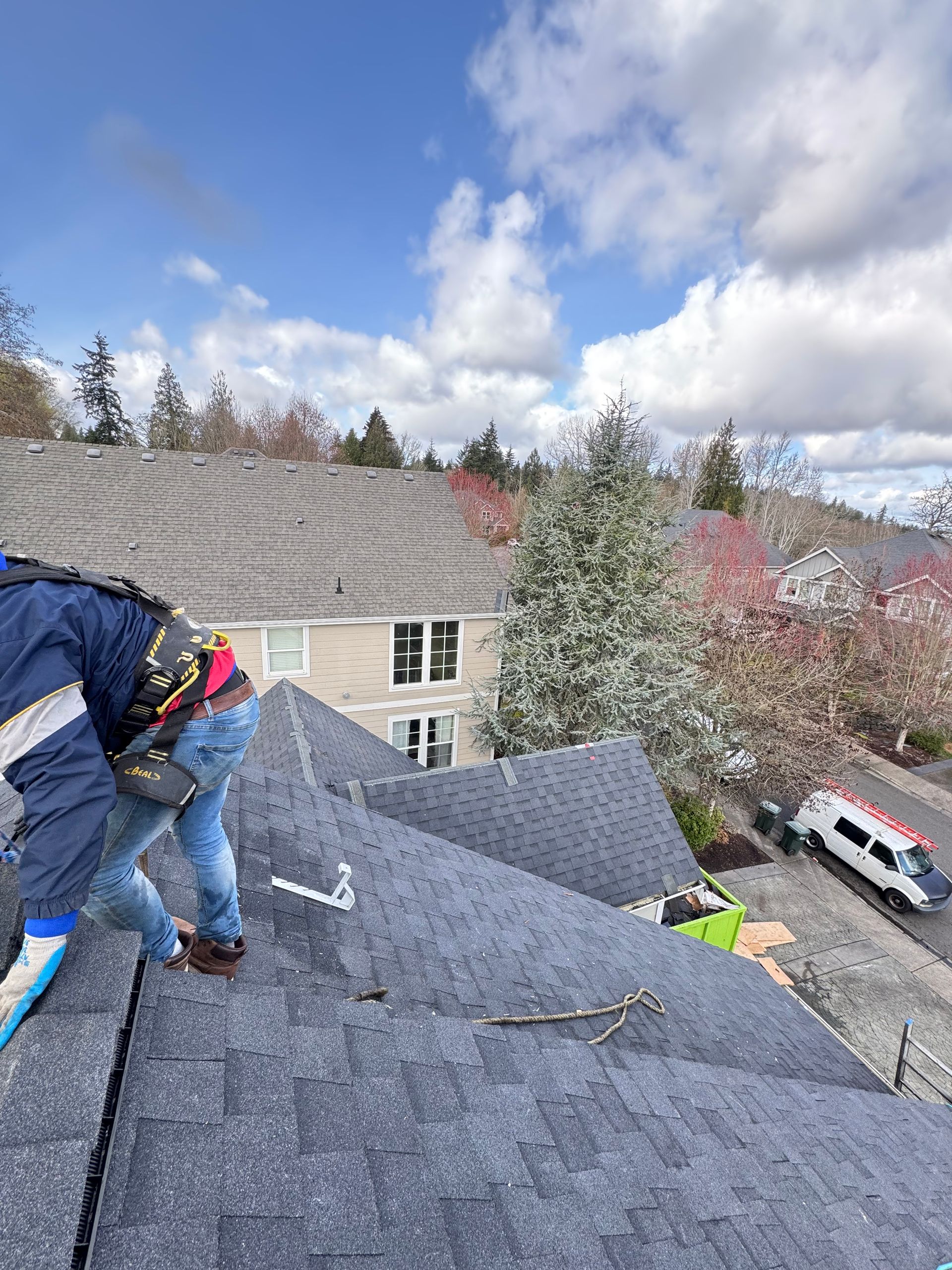 Worker on a steep shingled roof, installing shingles near trees and a residential building.