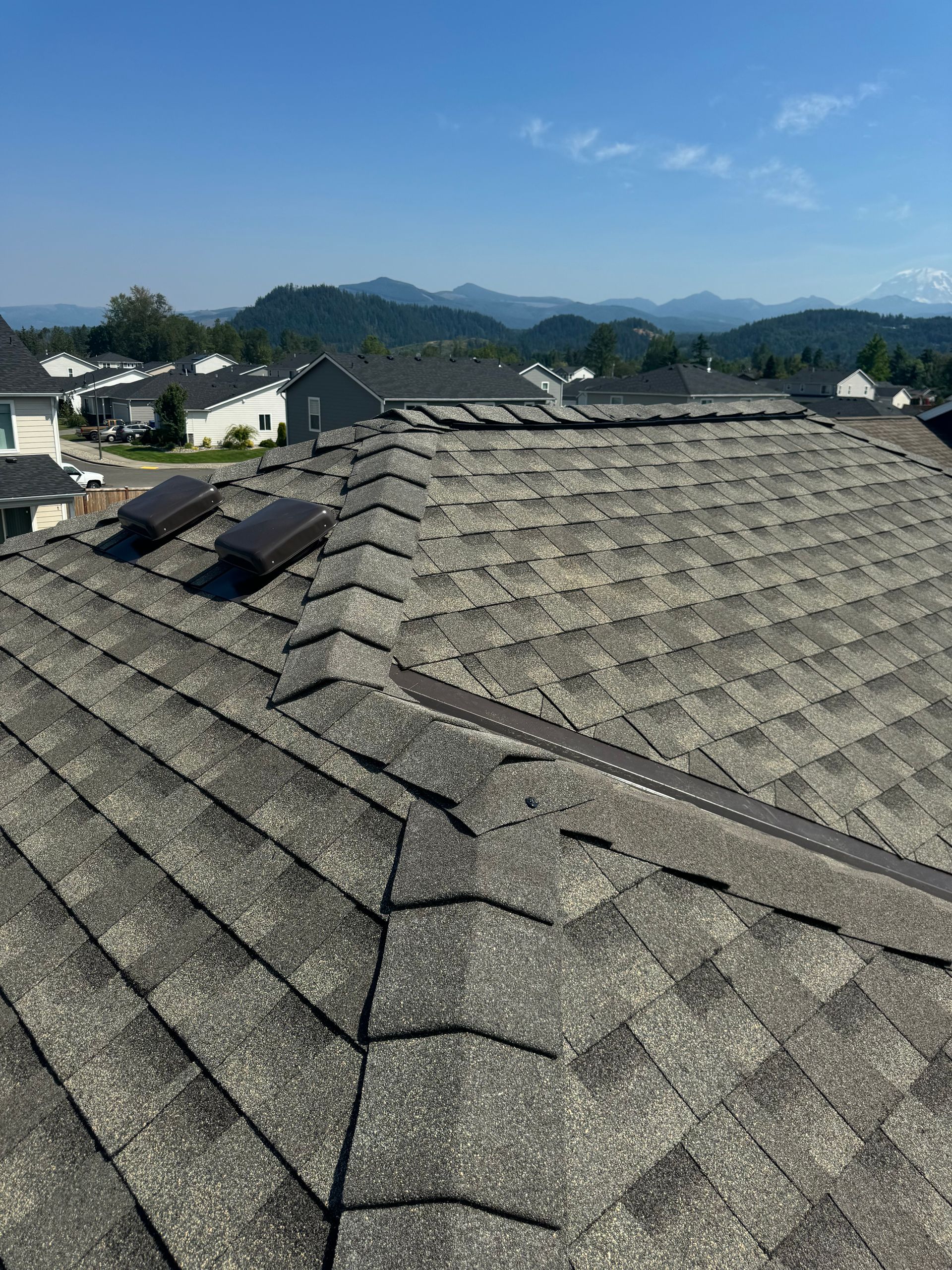 View from a gray shingled rooftop toward distant mountains and houses under a clear blue sky