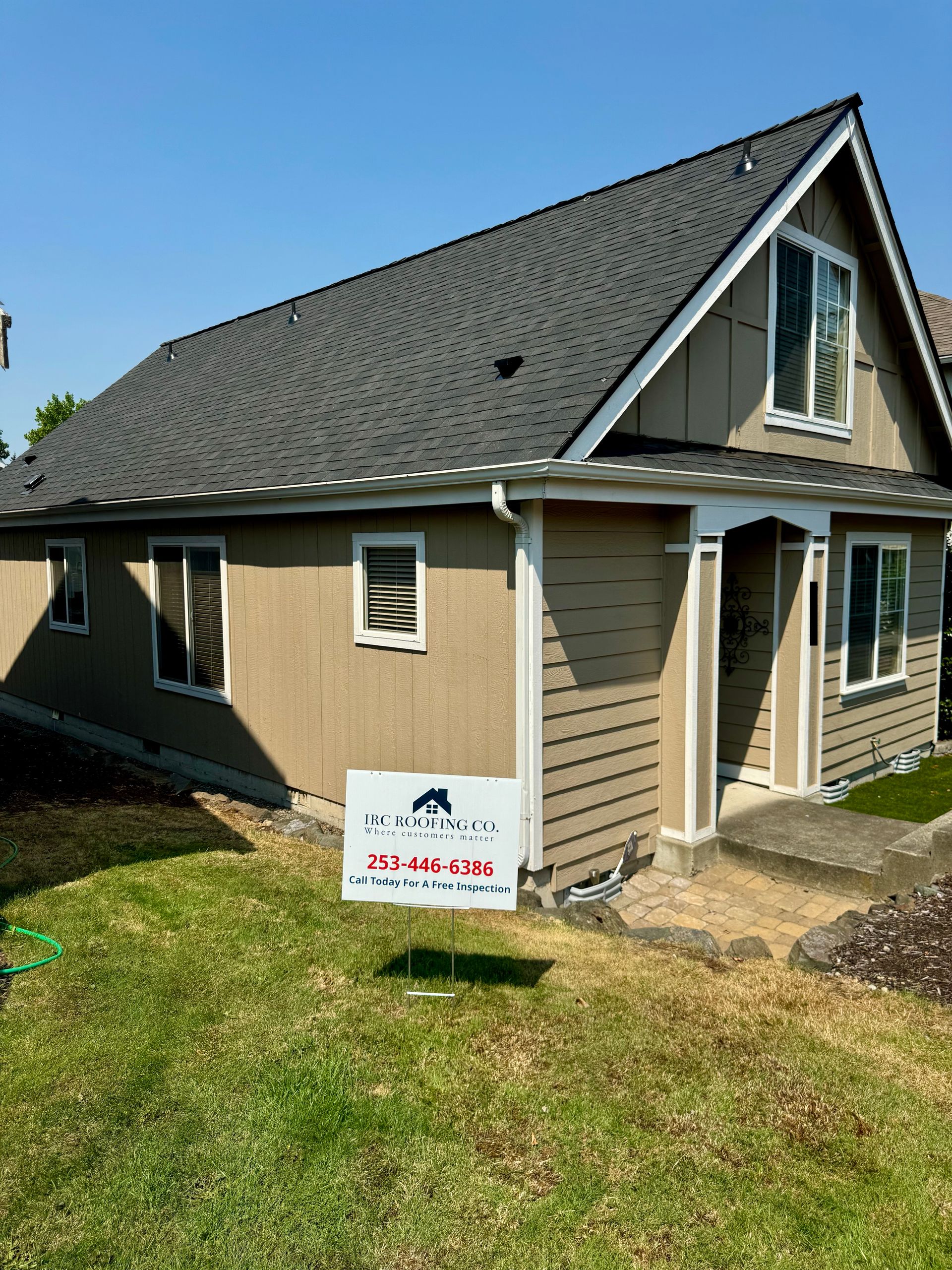 Tan house with steep roof and front porch in a sunny yard, with a small sign in the lawn.