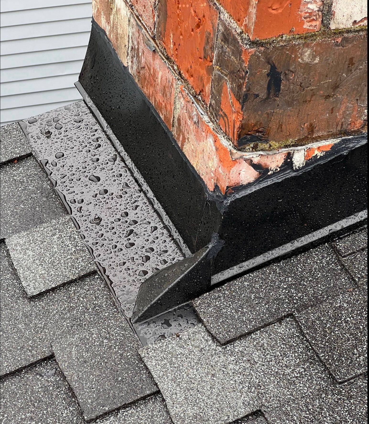 Damaged brick chimney with black flashing on a shingled roof, showing wear and rust stains.