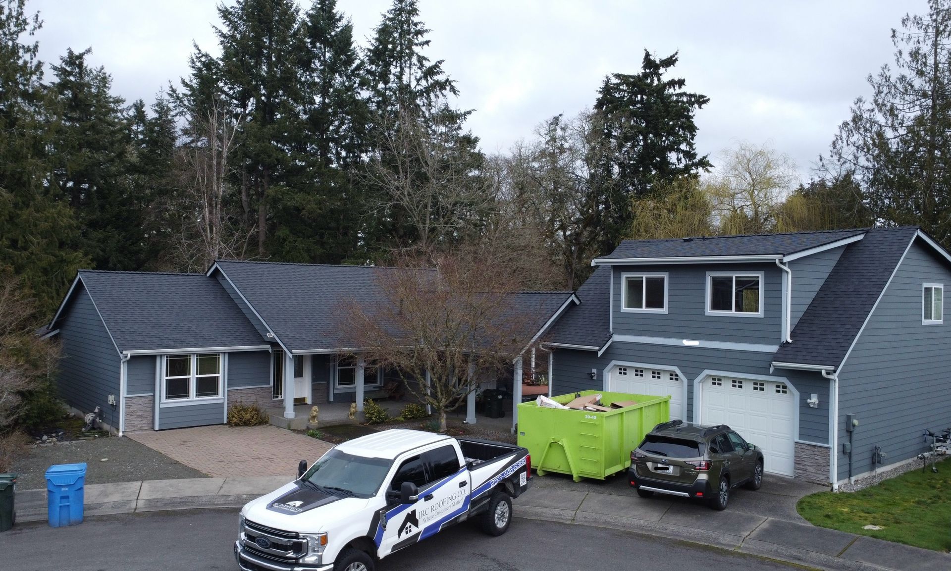 Suburban house with two cars in the driveway and a bright green dumpster beside the garage.