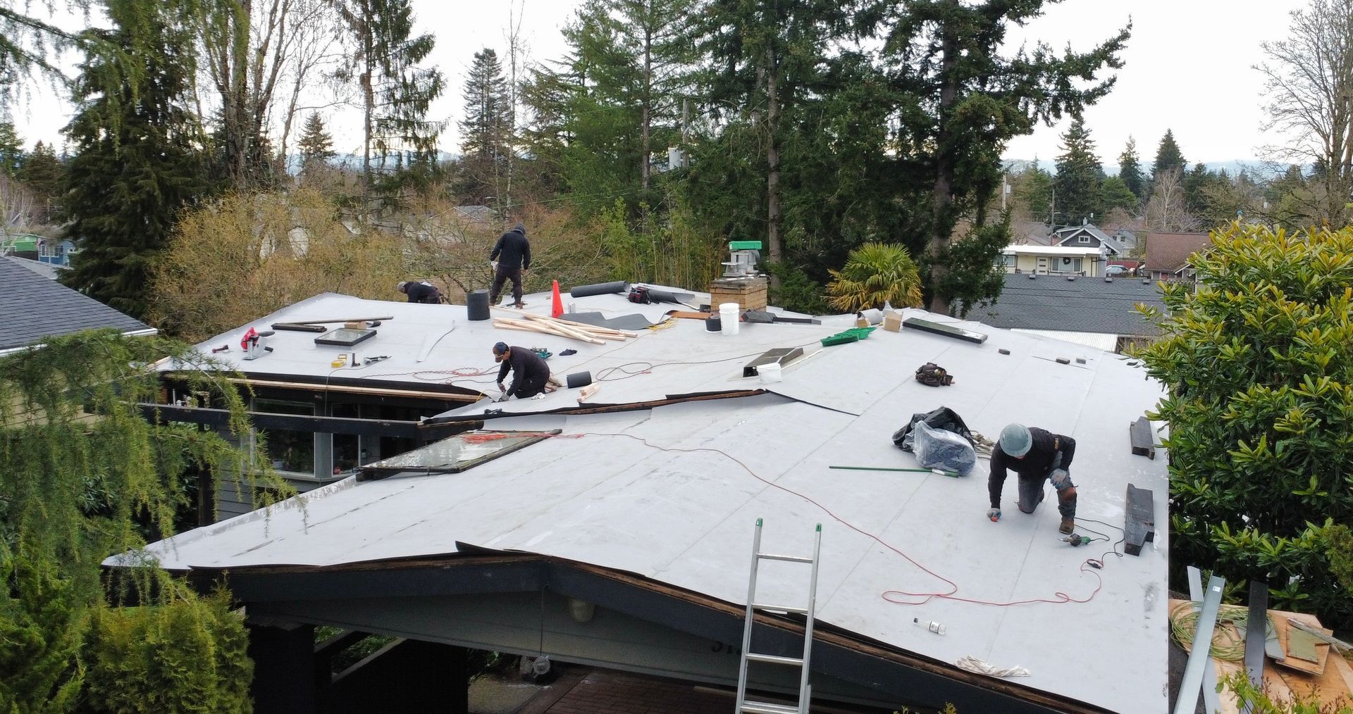 Roofers working on a large white roof in a wooded residential area, with ladders and tools visible.
