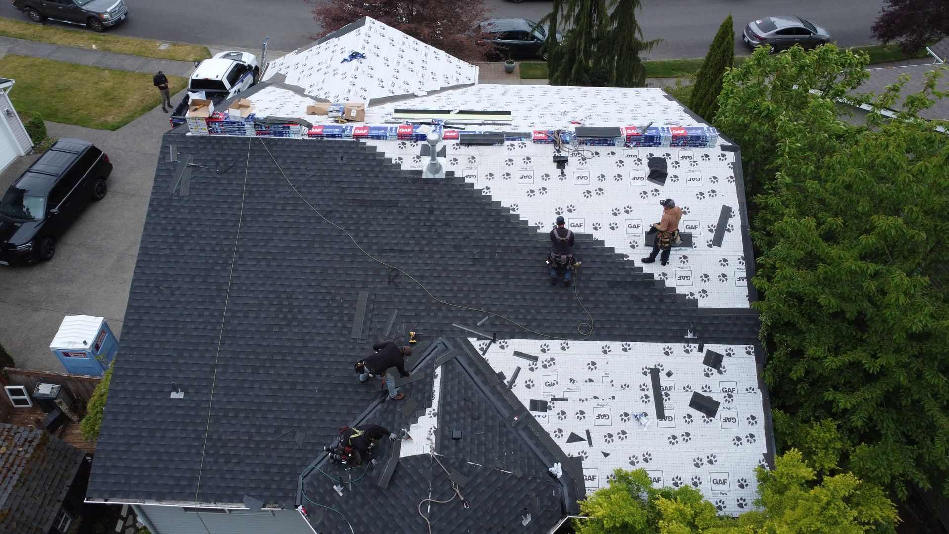 Aerial view of roofers repairing a house roof with dark shingles and white underlayment, surrounded by trees.