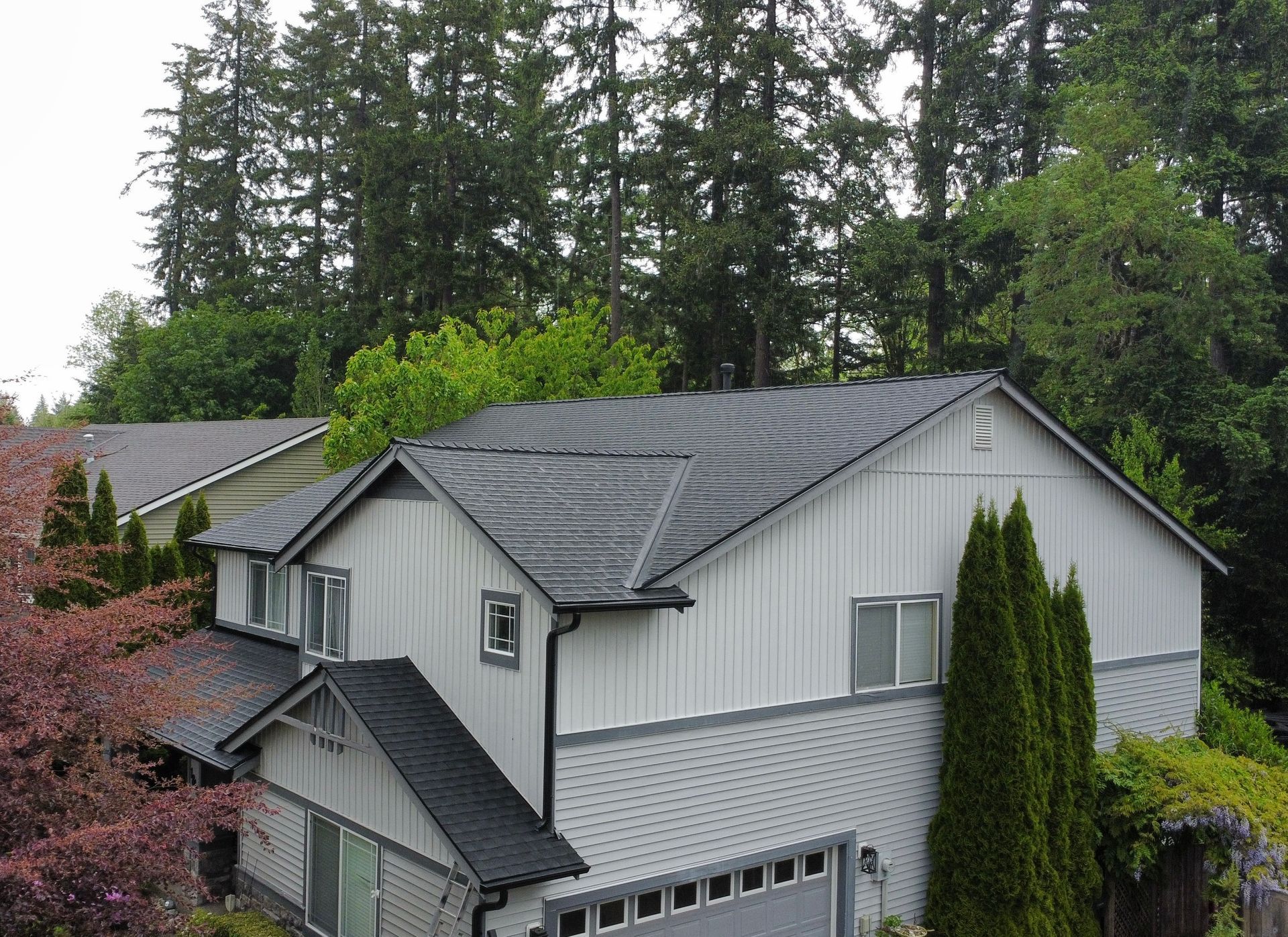 Gray house with dark roof and tall evergreens in a wooded suburban setting