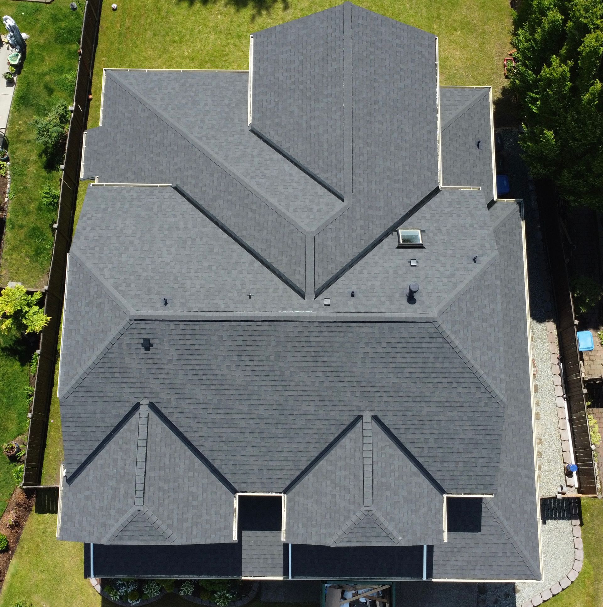 Aerial view of a gray-shingled house roof with multiple gables and vents, surrounded by greenery.