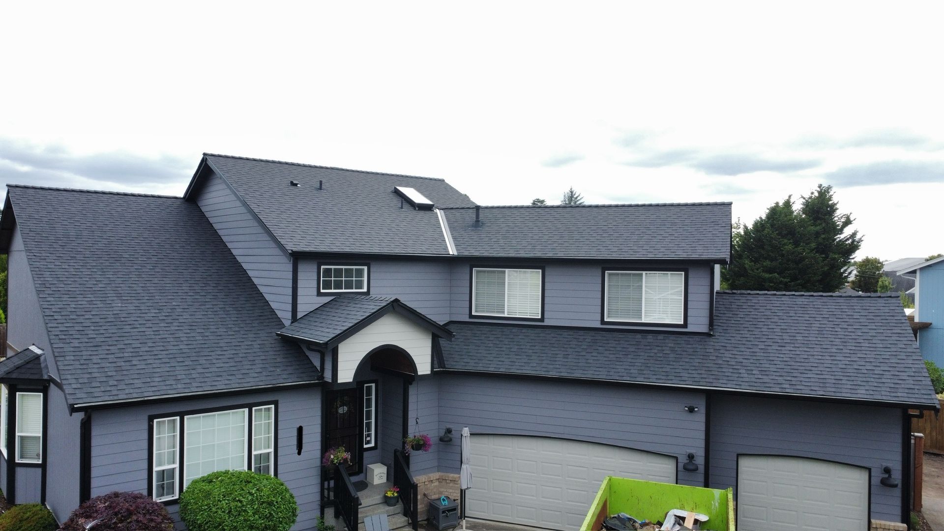 Two-story gray house with dark roof, front garage, and shrubs under a cloudy sky