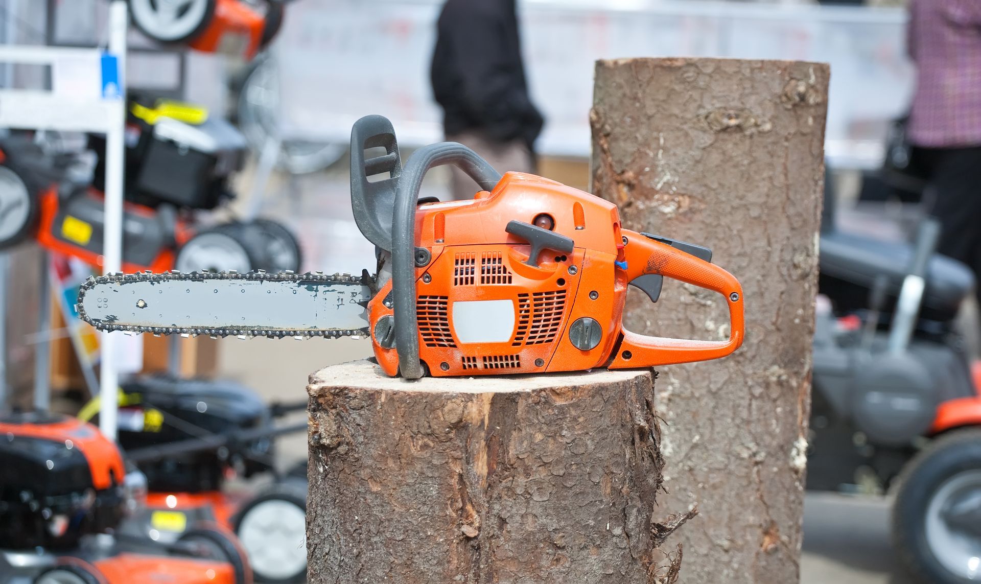 Orange chainsaw resting on a tree stump, with other lawn equipment in background.