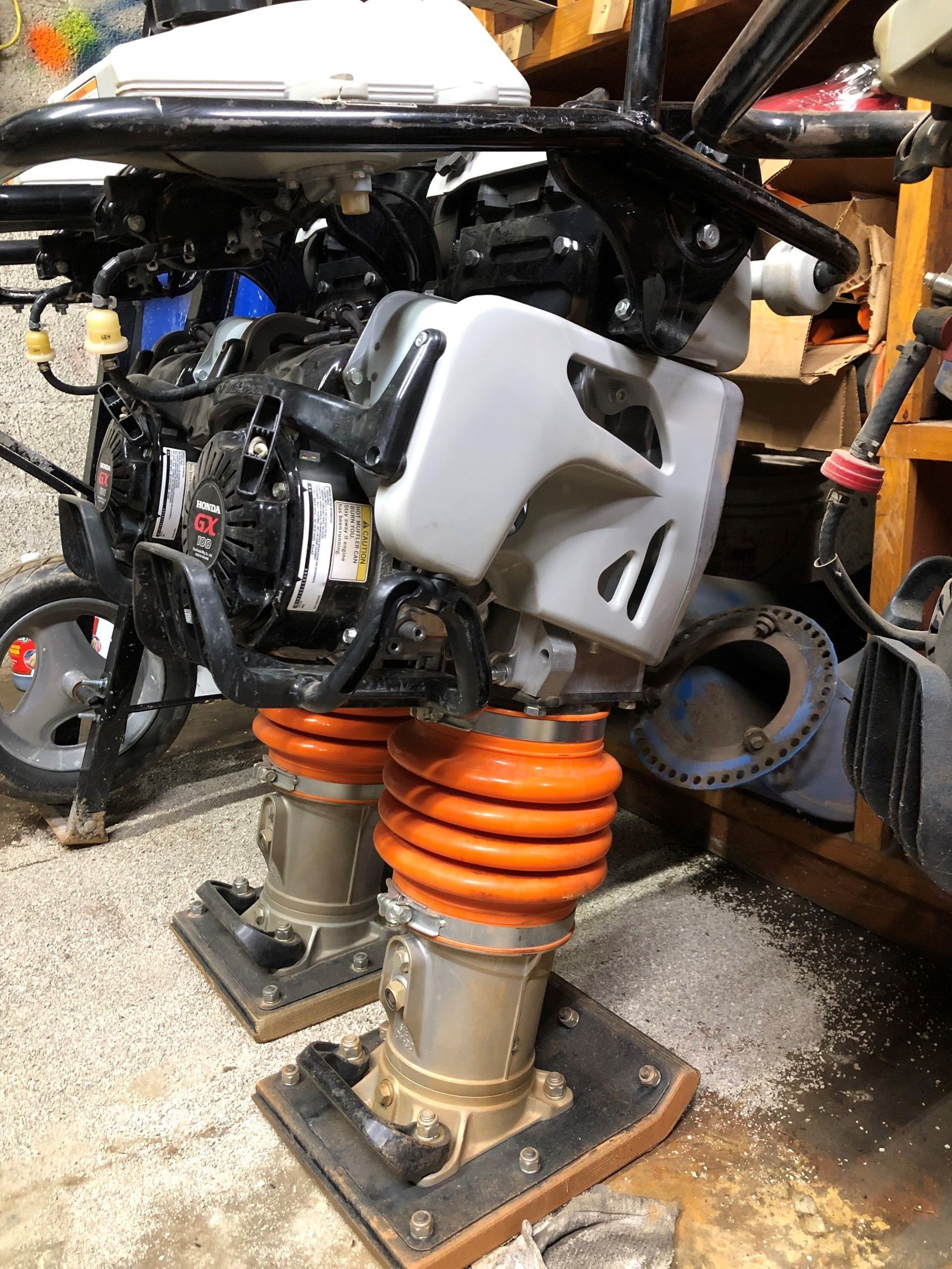 A jumping jack compactor in a workshop. Gray engine cover, orange bellows, dark frame and handle.