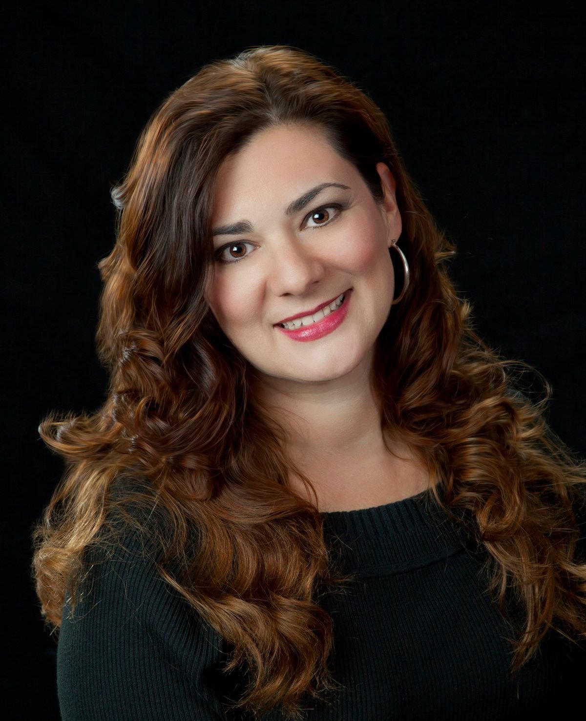Dr. Janee Atkinson with long, curly brown hair smiles, wearing a black sweater against a dark background.
