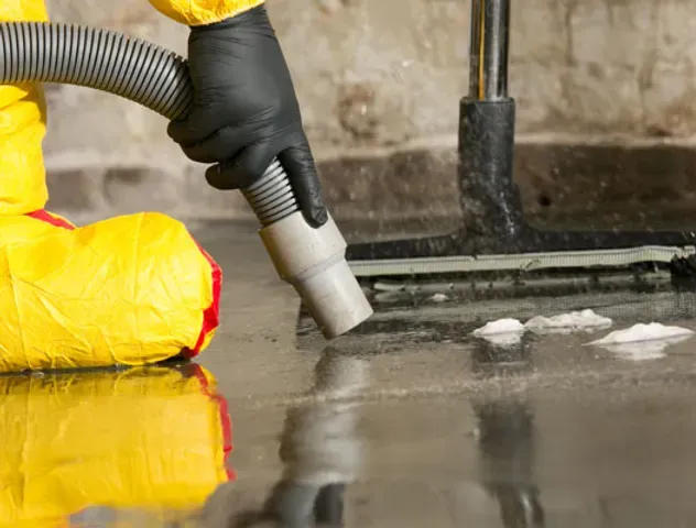 A person in a yellow suit is using a vacuum cleaner to clean a flooded basement.