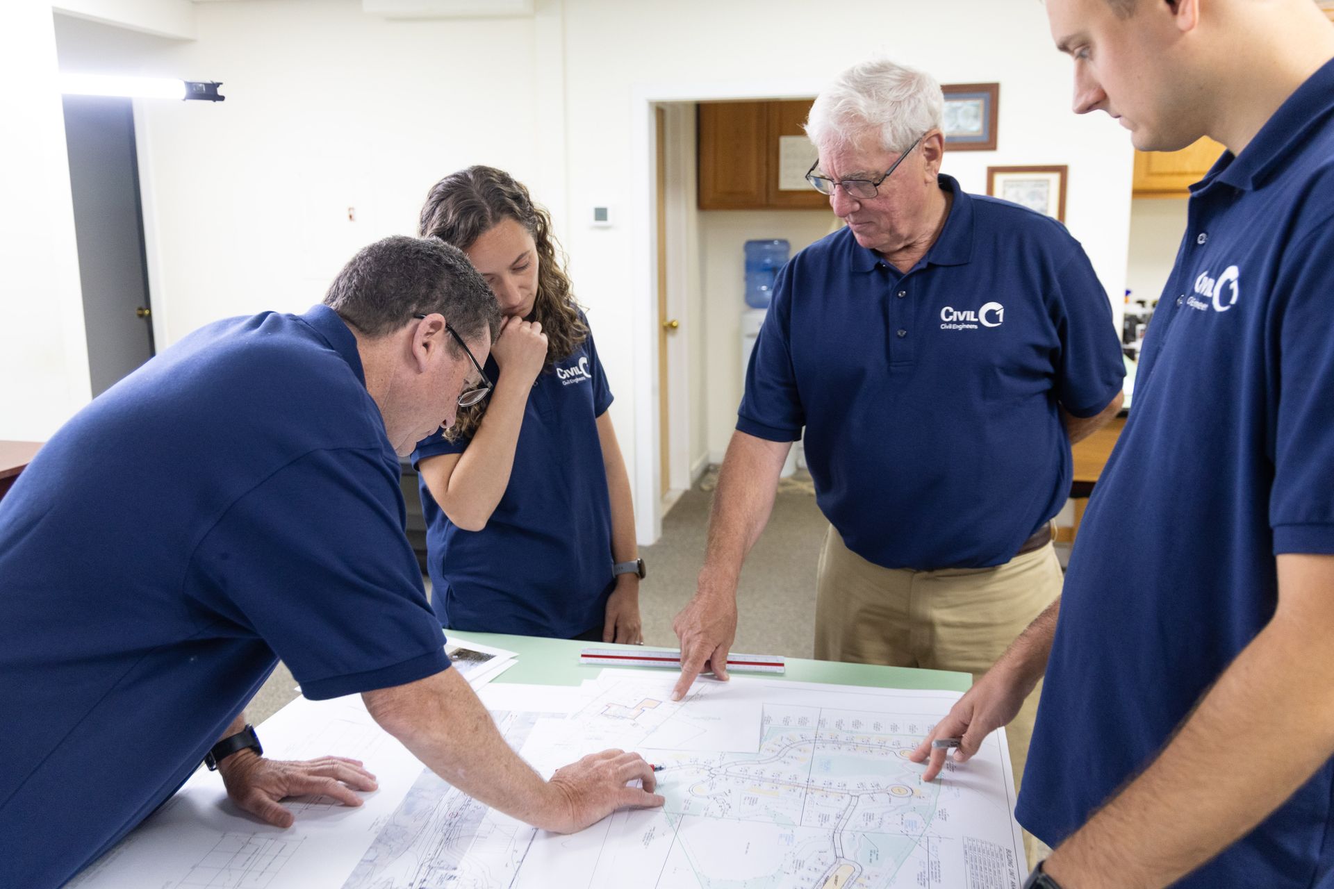 Four people in blue shirts examine a map spread on a table in a room.