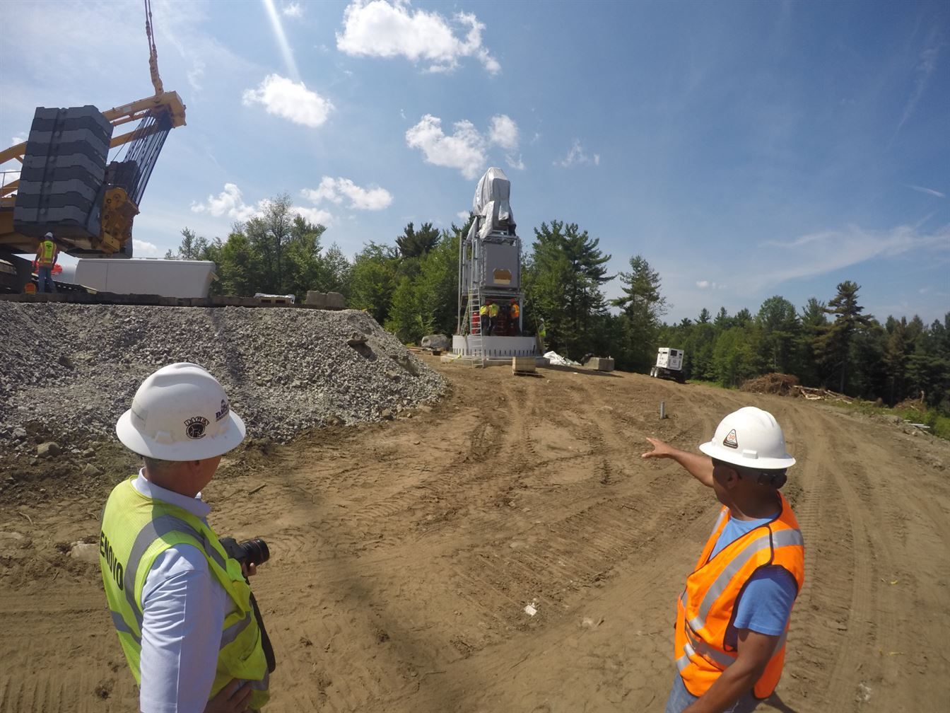 Two engineers are standing in a dirt field looking at a wind turbine being constructed.
