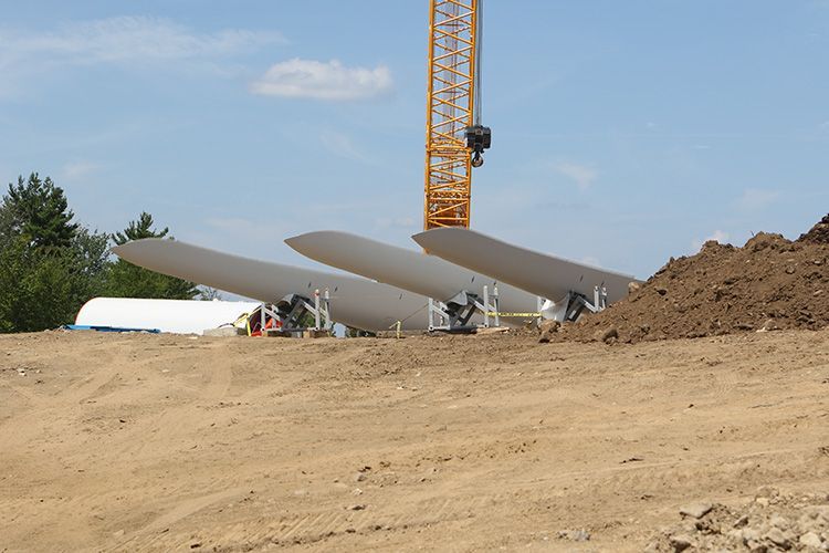 Three wind turbine blades are sitting in a dirt field with a crane in the background