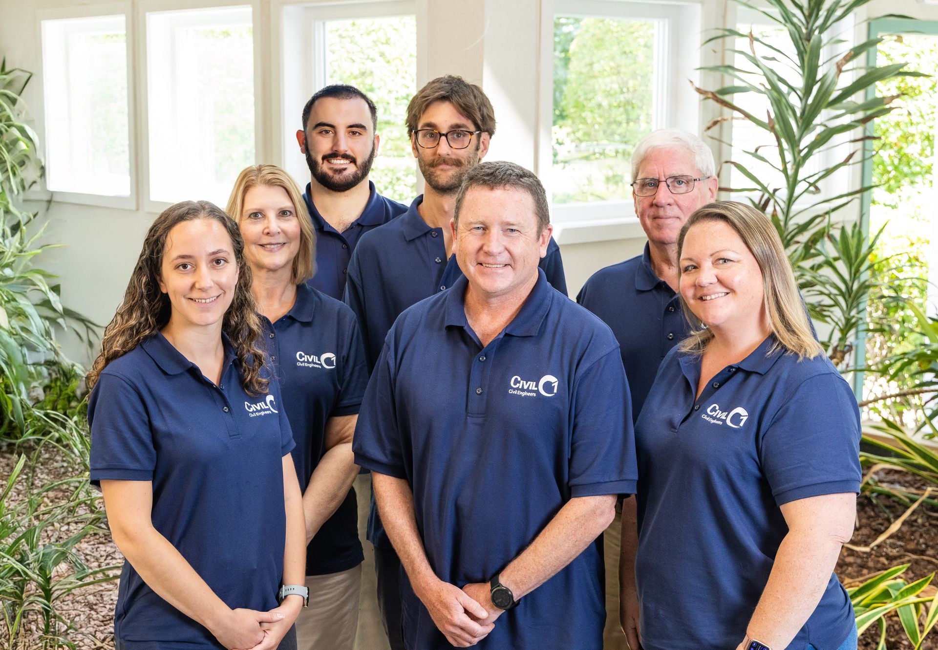 Group of seven people wearing blue shirts, smiling in an office setting with plants.
