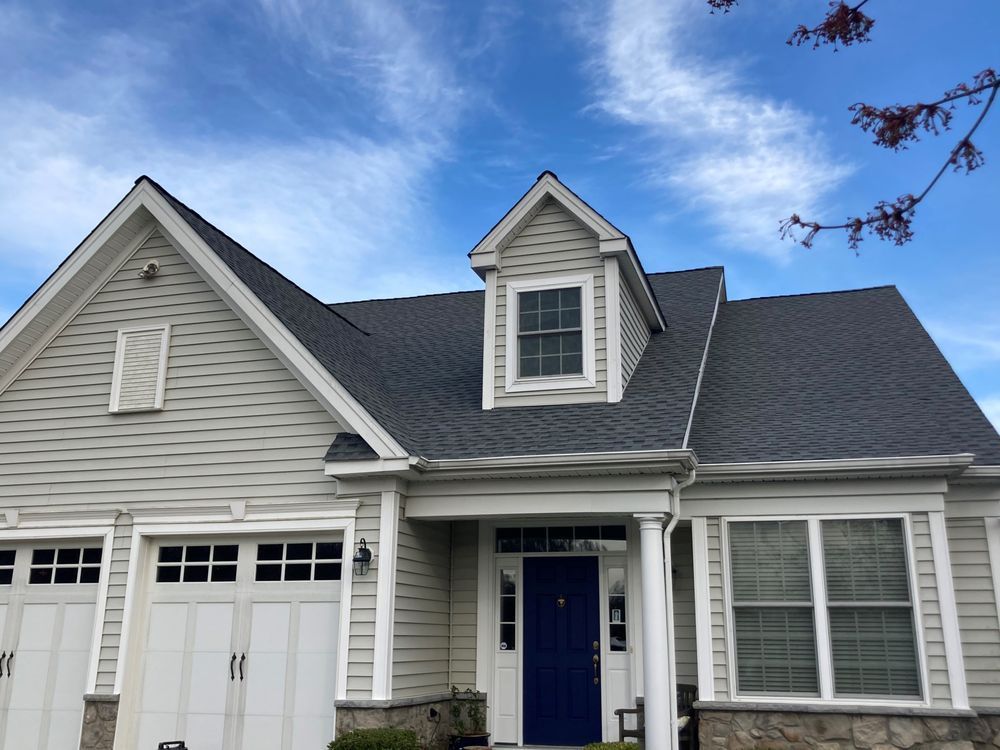 Gray house with white trim, blue front door, and dormer windows under a partly cloudy sky