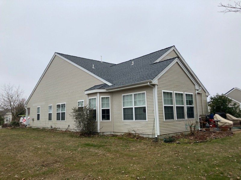 Suburban beige house with gray roof and white trim on a grassy lot under a cloudy sky