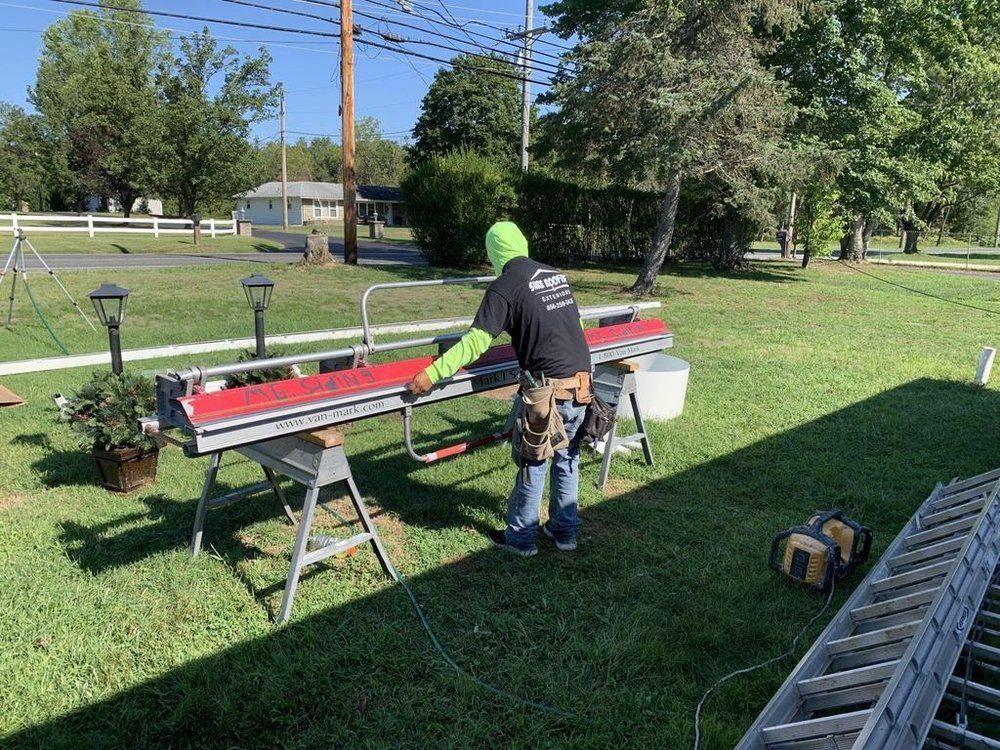 Worker in a green helmet measuring red rails on sawhorses in a sunny yard with tools and a ladder nearby.