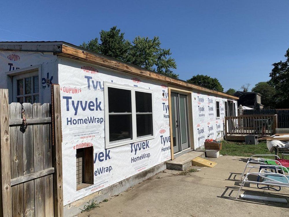 Small shed under construction, wrapped in Tyvek with unfinished window and wooden deck outside.
