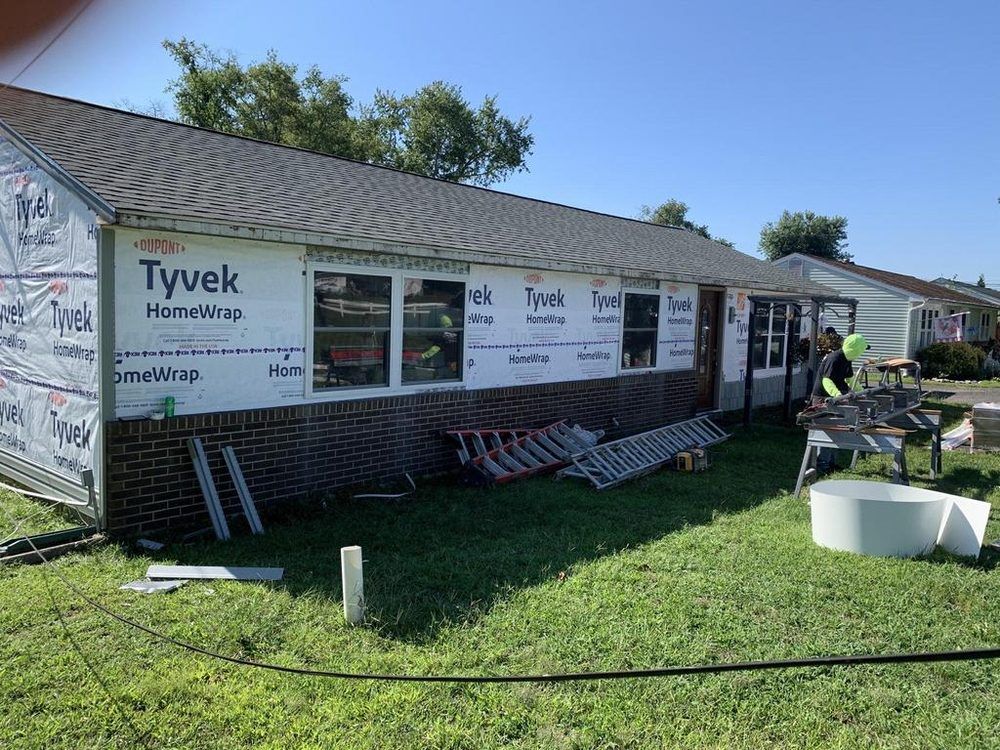 House under construction wrapped in Tyvek, with ladders, tools, and workers on a sunny lawn