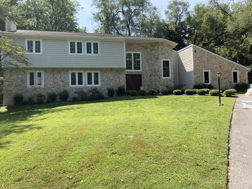 Two-story stone house with a green lawn and a driveway under bright sunlight.
