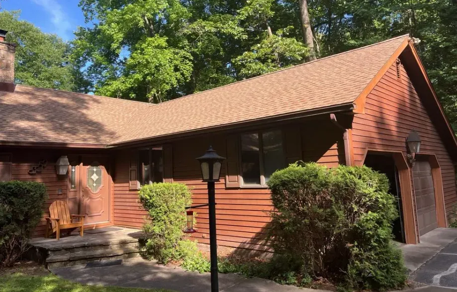 Red wooden house with a sloped roof, front door, shrubs, and a lamppost in a leafy yard