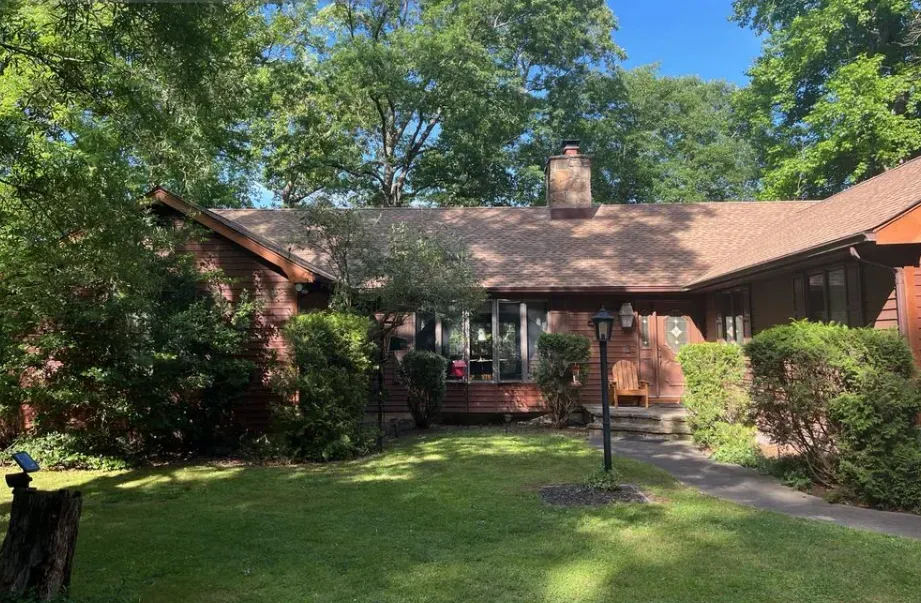Brick house with a red roof, surrounded by trees and bushes, viewed from a green front lawn.