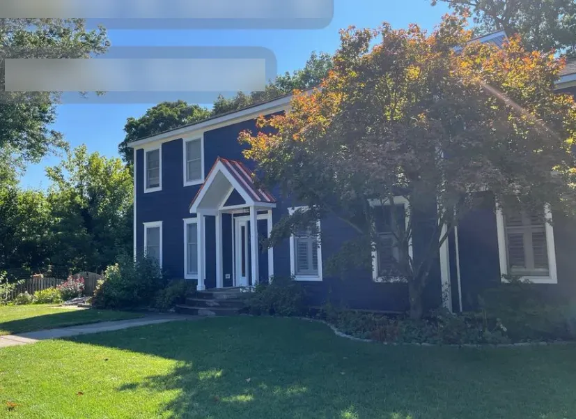 Blue two-story house with white trim and a large leafy tree in the front yard under a sunny sky