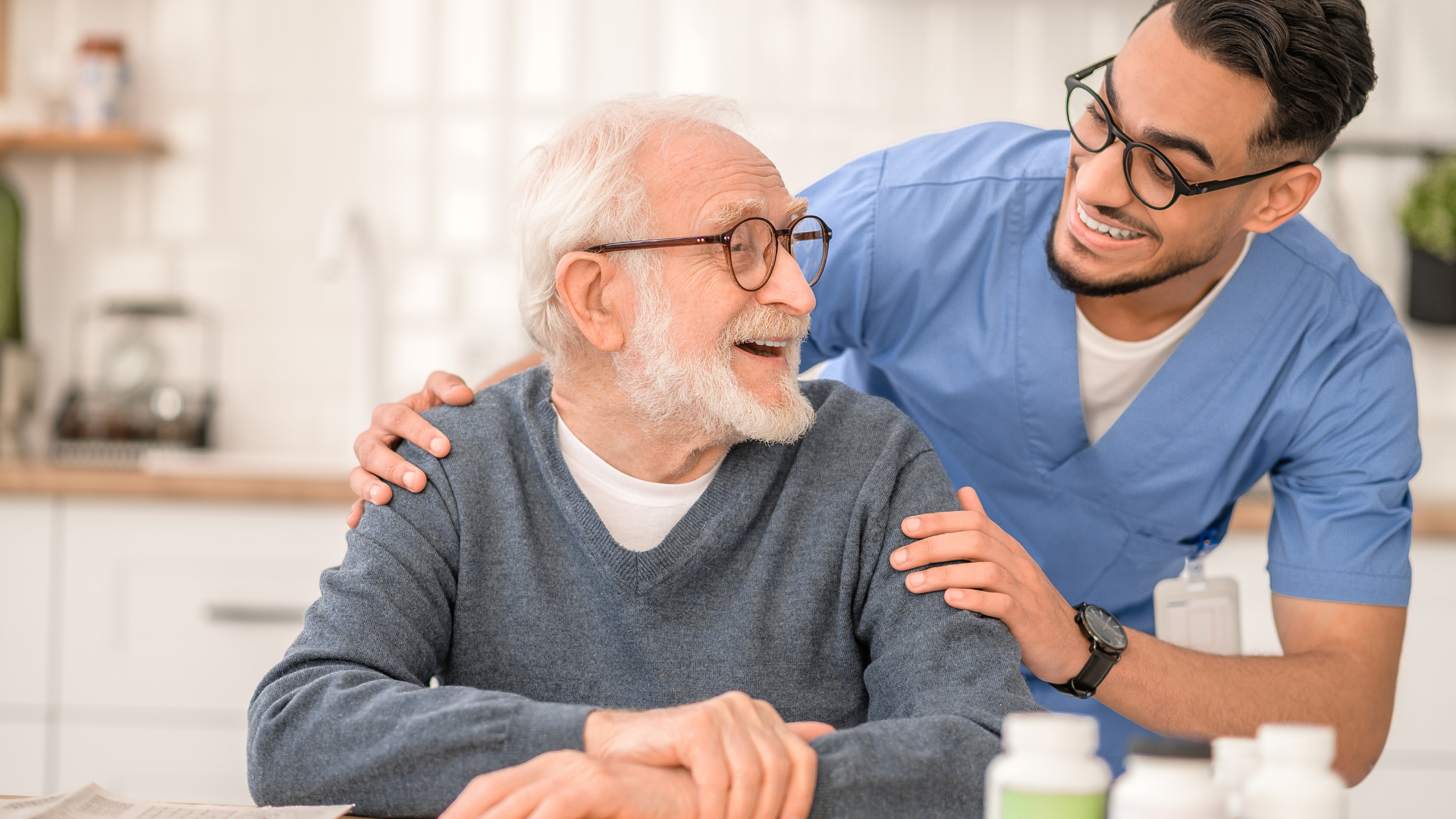 Caregiver in blue smiles and supports an older man seated at a table in a bright home setting