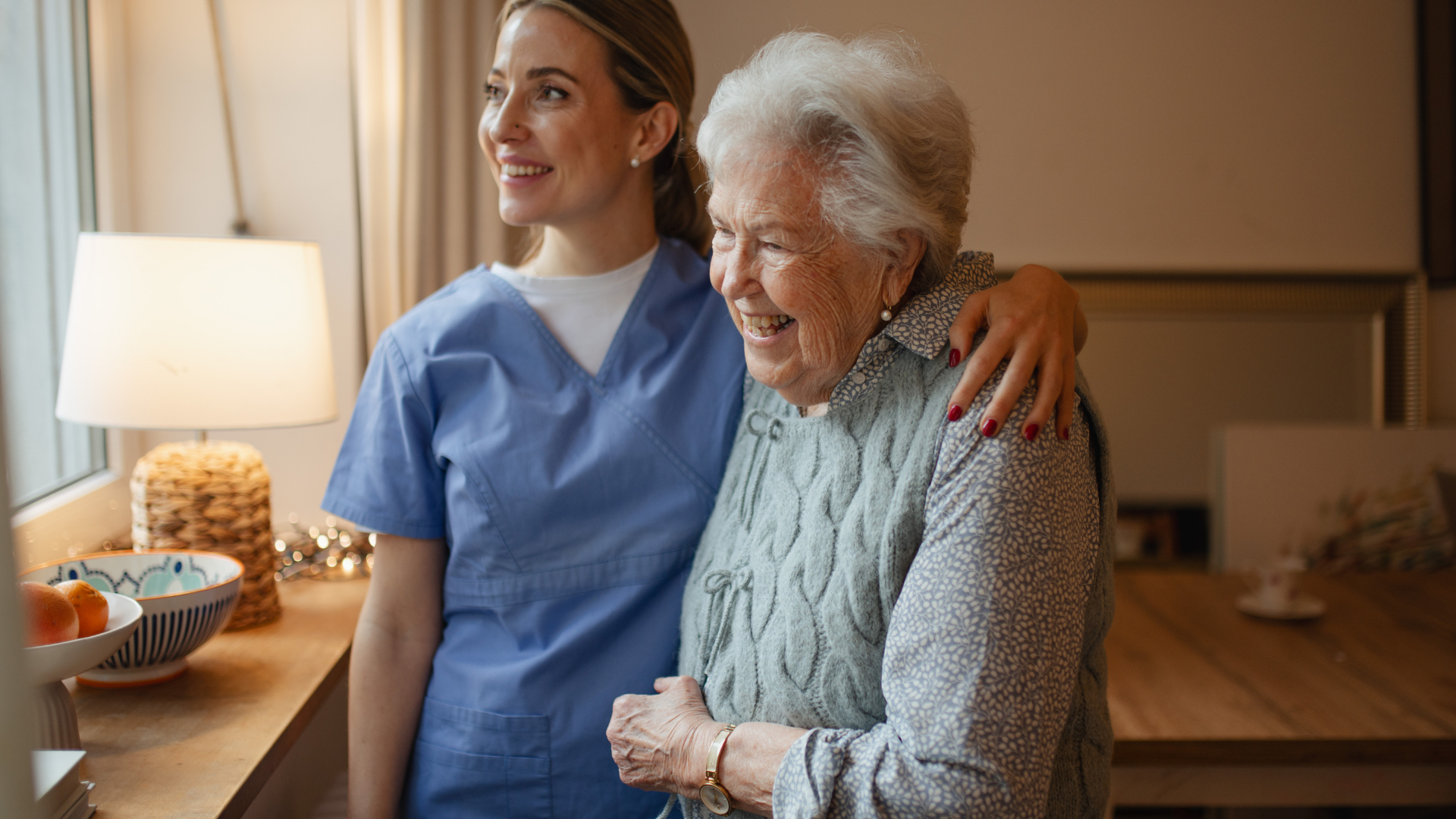 Caregiver with an elderly woman standing together in a softly lit home room, both smiling warmly.