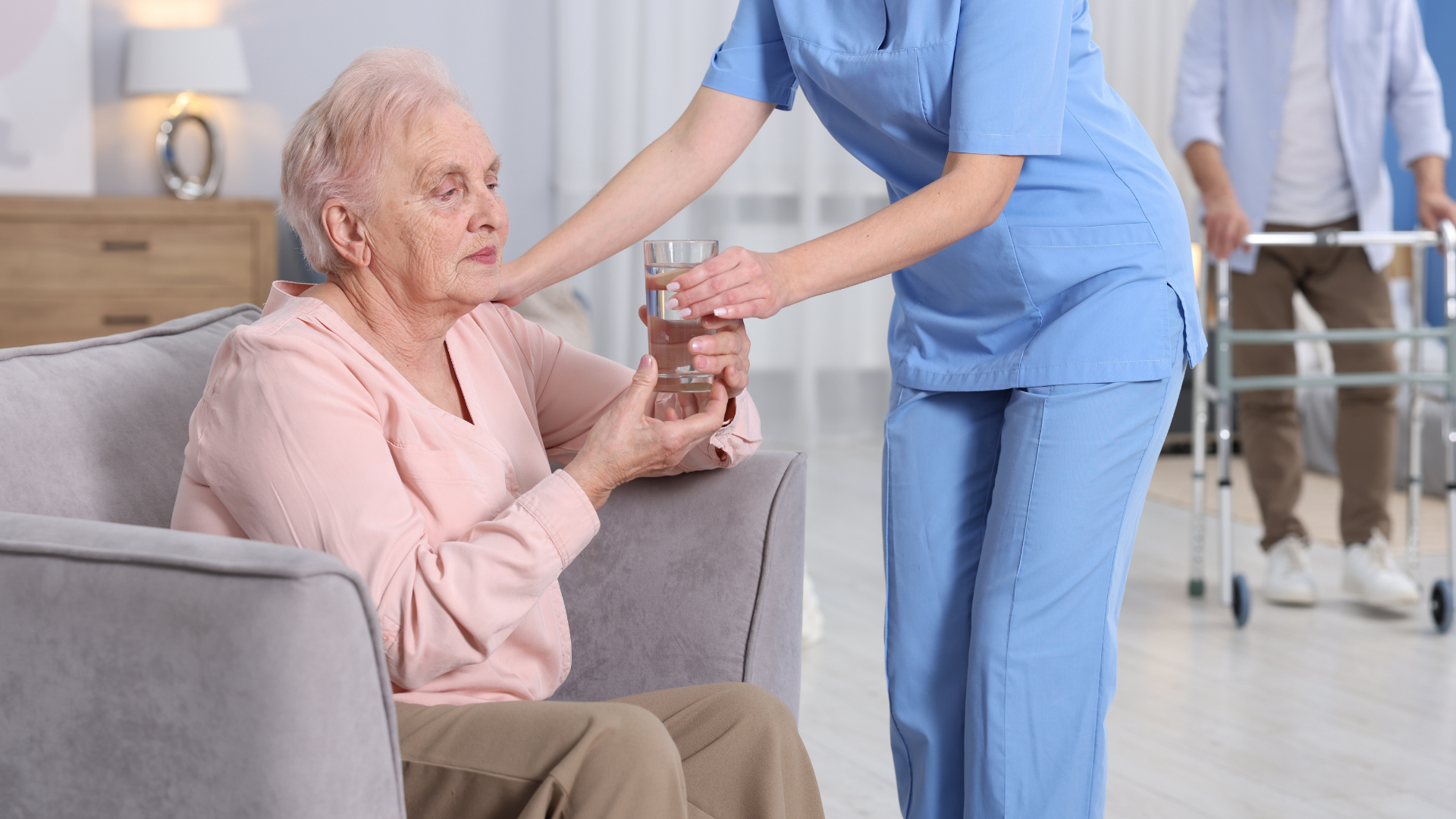 Caregiver in blue assists seated patient in a bright hospital lounge; another staff member walks in background.
