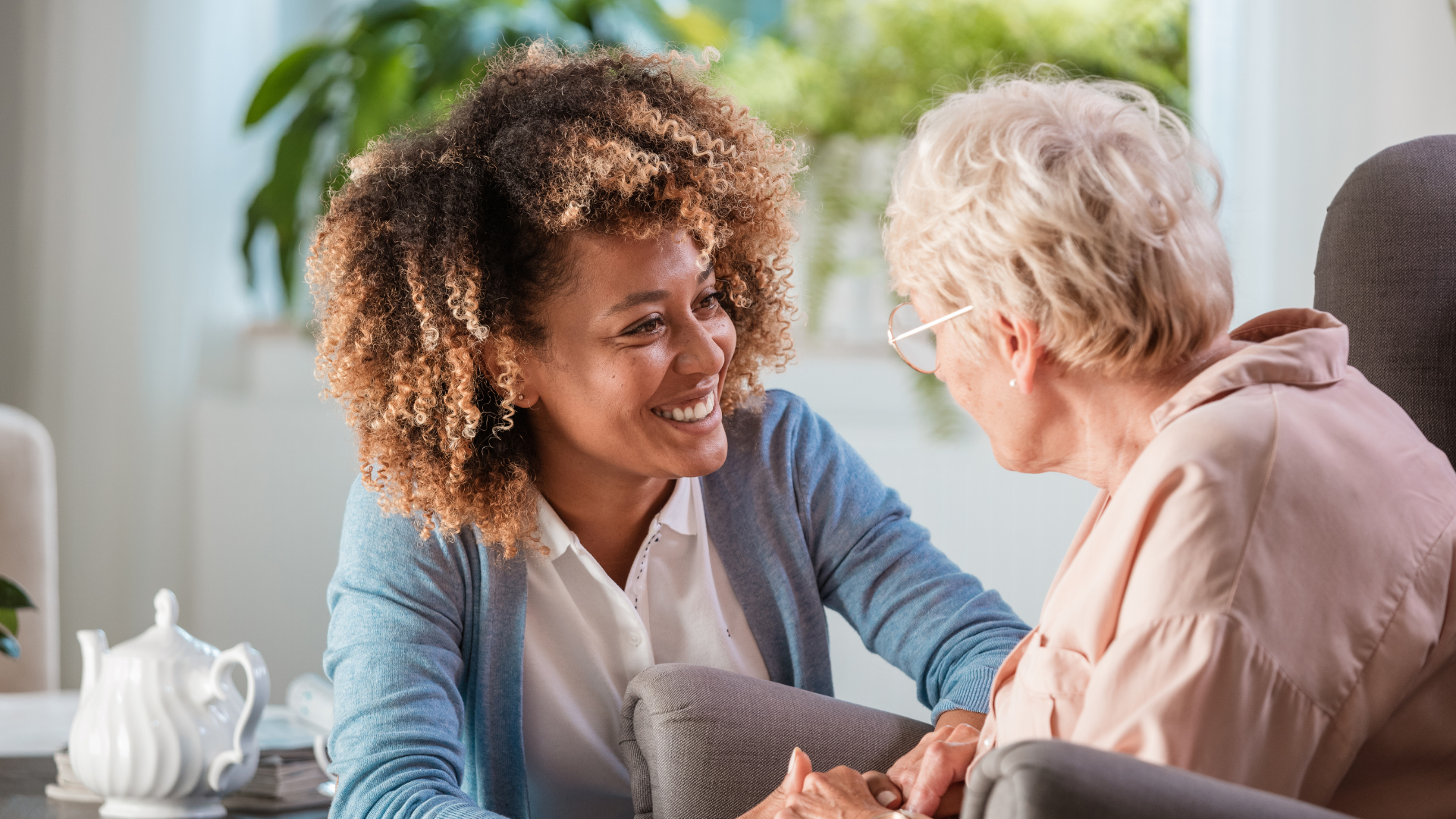 Caregiver helping an elderly person stand in a living room near a sofa and dresser