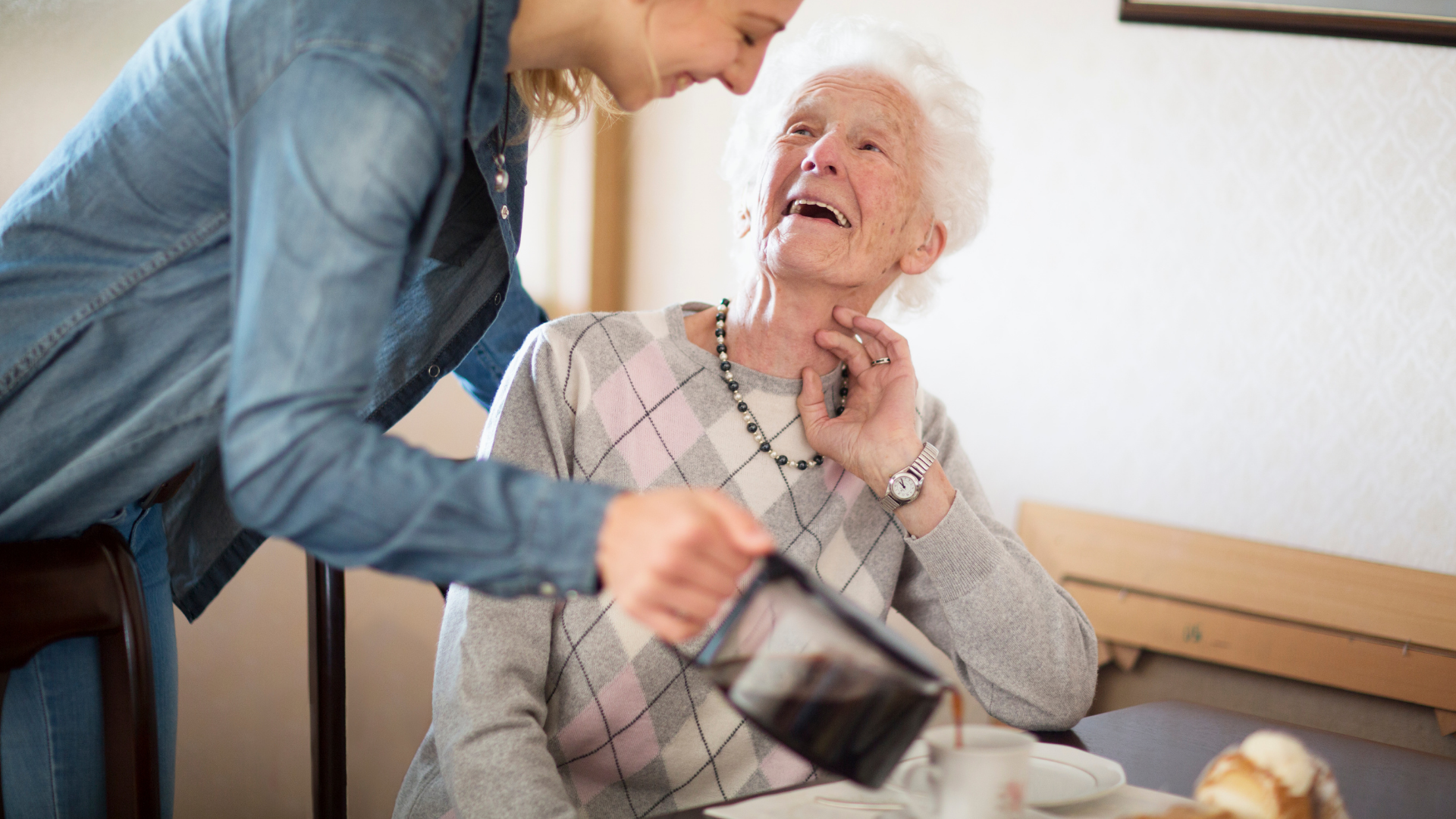 Caregiver in blue shirt helping an elderly woman in a bed or chair, with a warm, attentive expression.
