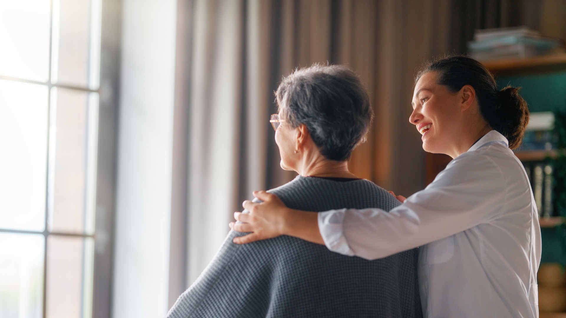 Two people by a window, one gently consoling the other with an arm around their shoulder.