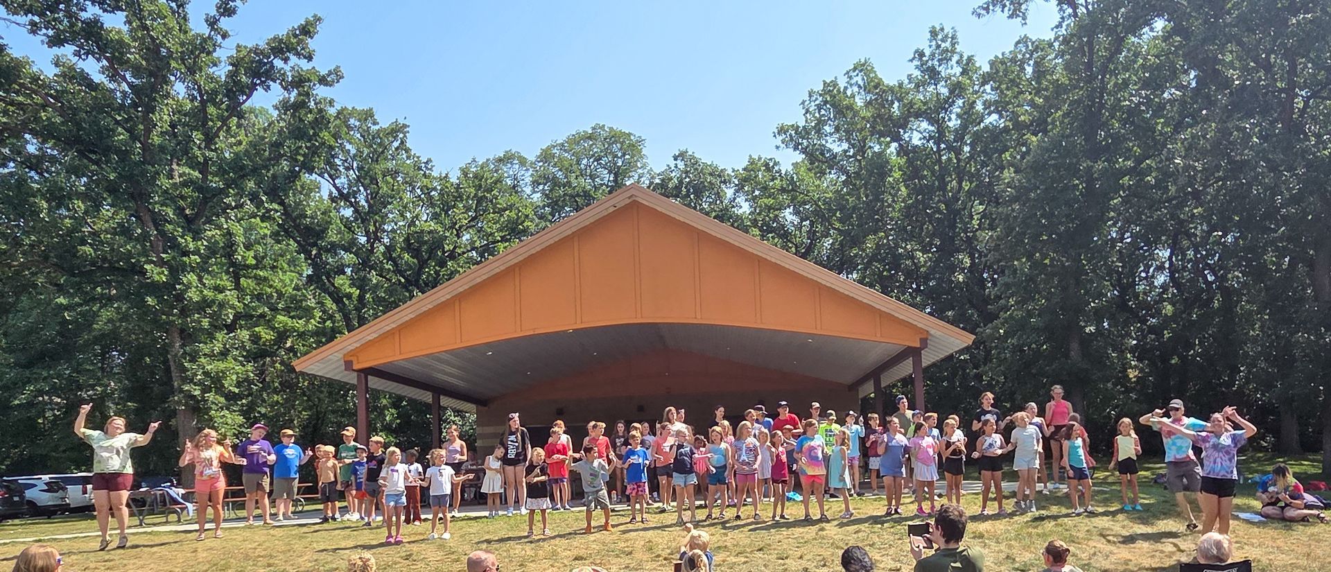 Group of children standing in a line under a covered structure, some clapping.