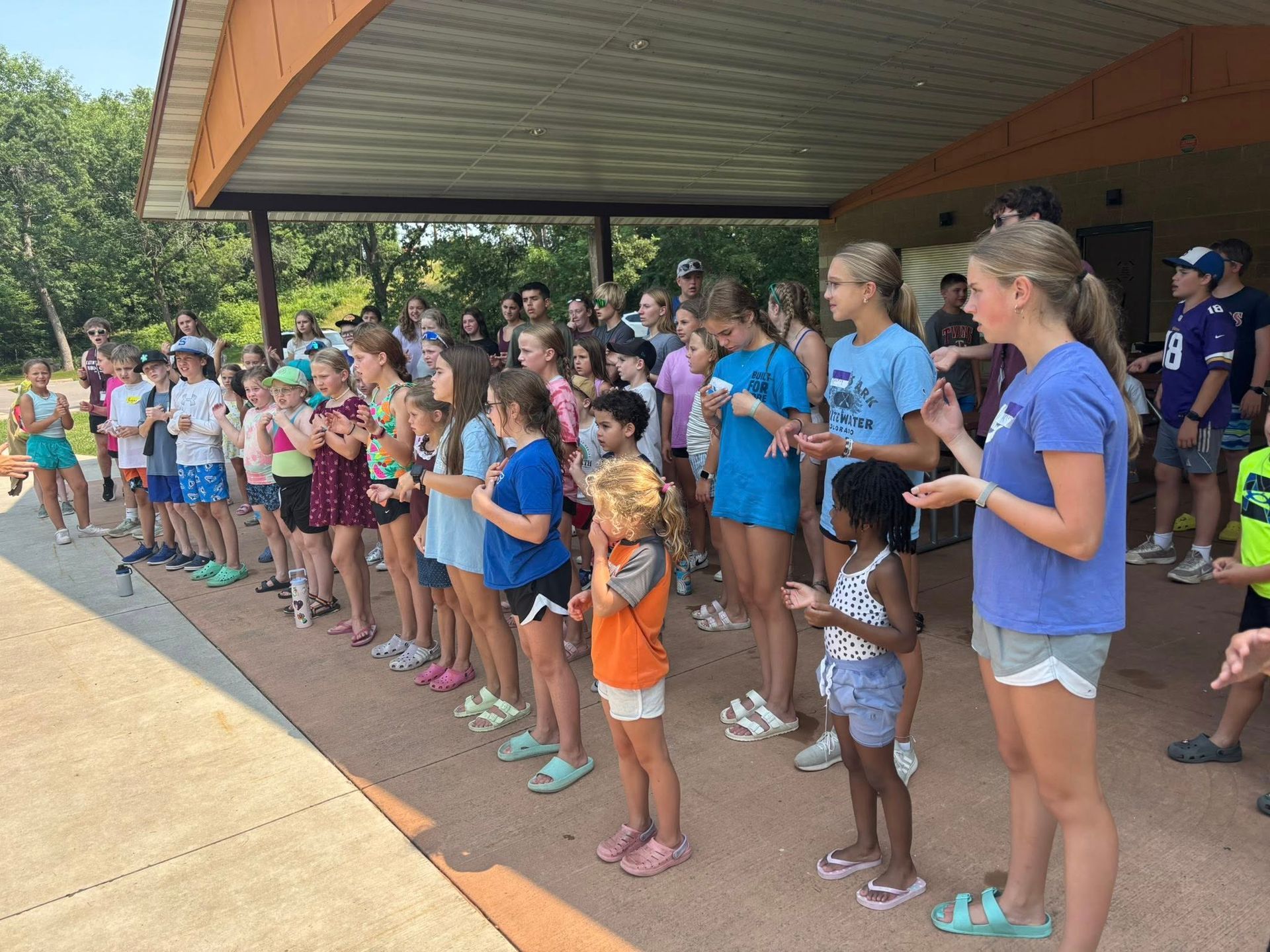 Group of children standing in a line under a covered structure, some clapping.