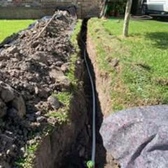 Trenches beside a grassy yard with exposed black pipe and piles of dirt from excavation
