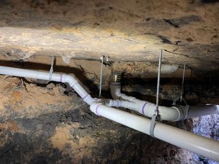 Exposed white PVC pipes and hanging cables under a rough dirt crawlspace ceiling