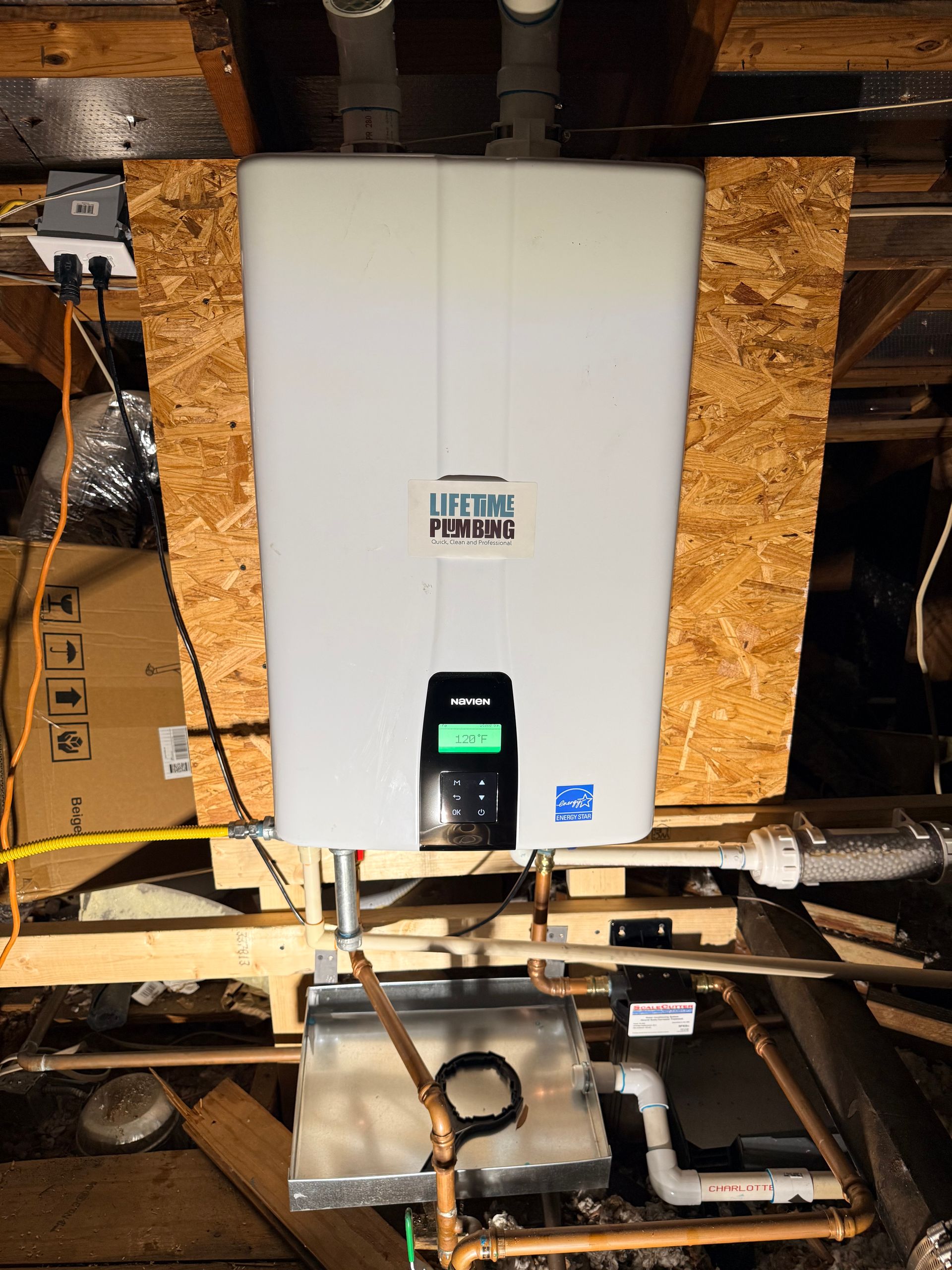 White utility boiler mounted in an unfinished attic with exposed wood framing and pipes