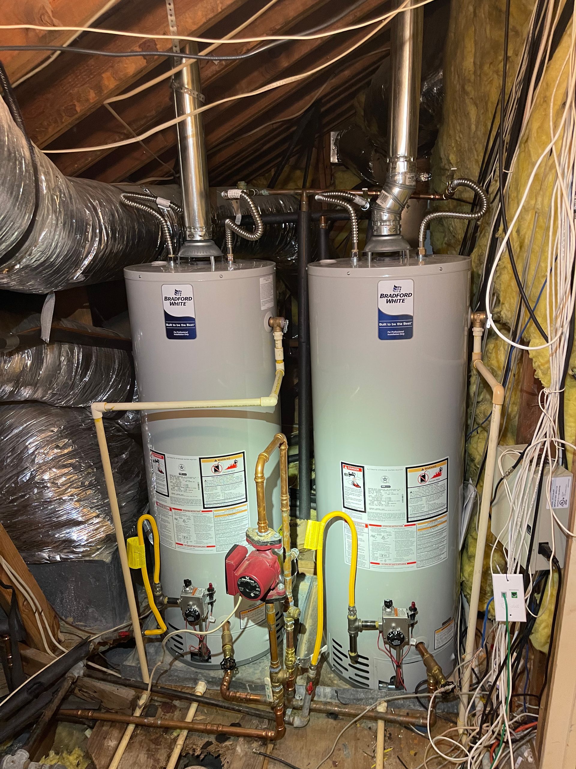 Boiler and hot water tank in a utility room with exposed pipes and heating controls on a white wood wall