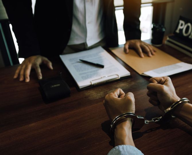 Person in handcuffs at a desk with another person, documents, and POLICE sign.